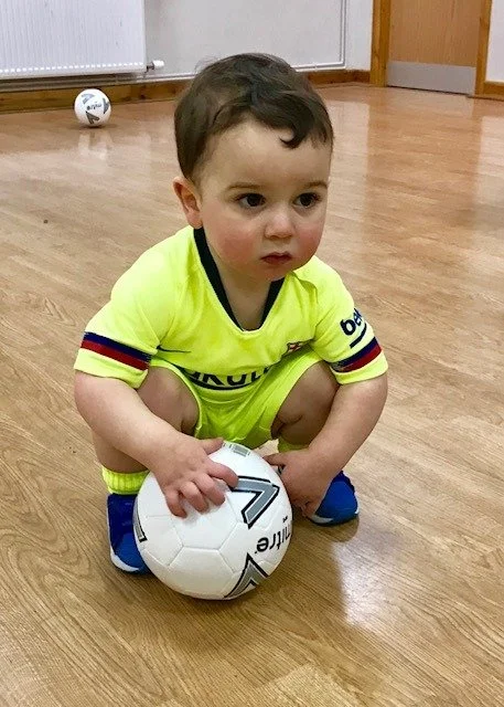 Young boy wearing a yellow sports jersey squatting on a wooden floor, holding a soccer ball. There is a small white ball nearby in the background.