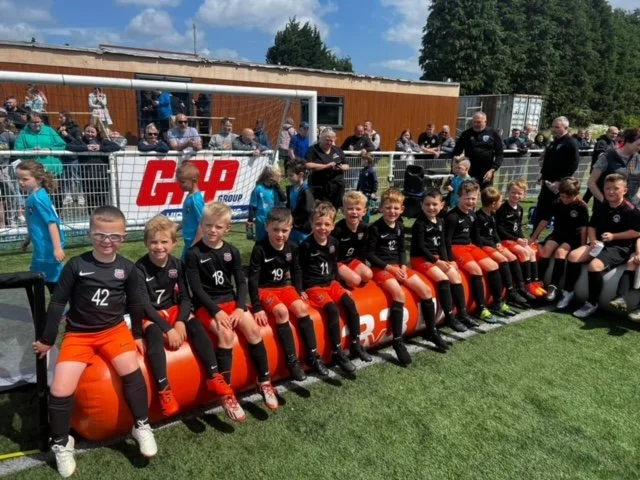 Youth soccer team sitting on a bench with their coach on a field with spectators behind a fence.