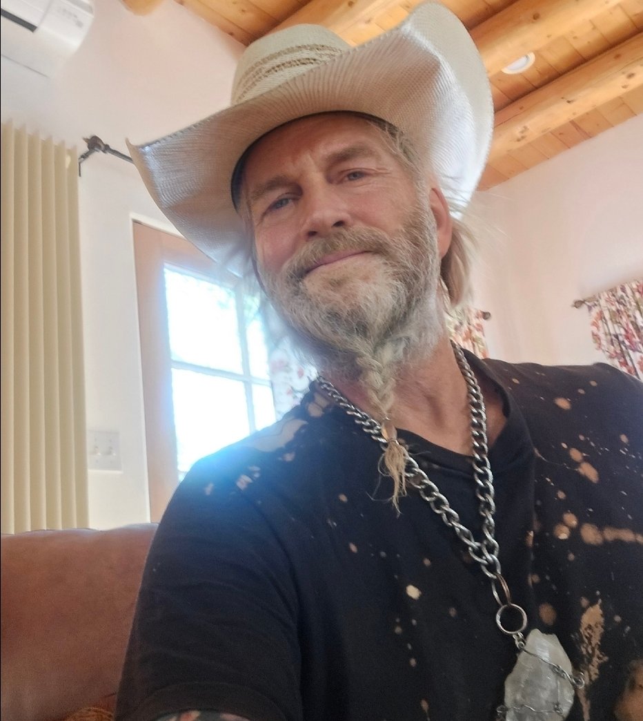 A man with a gray beard and long gray hair, wearing a white cowboy hat, a black distressed t-shirt, and a large chain necklace with a big crystal pendant, smiling at the camera inside a wooden ceiling room with floral curtains.