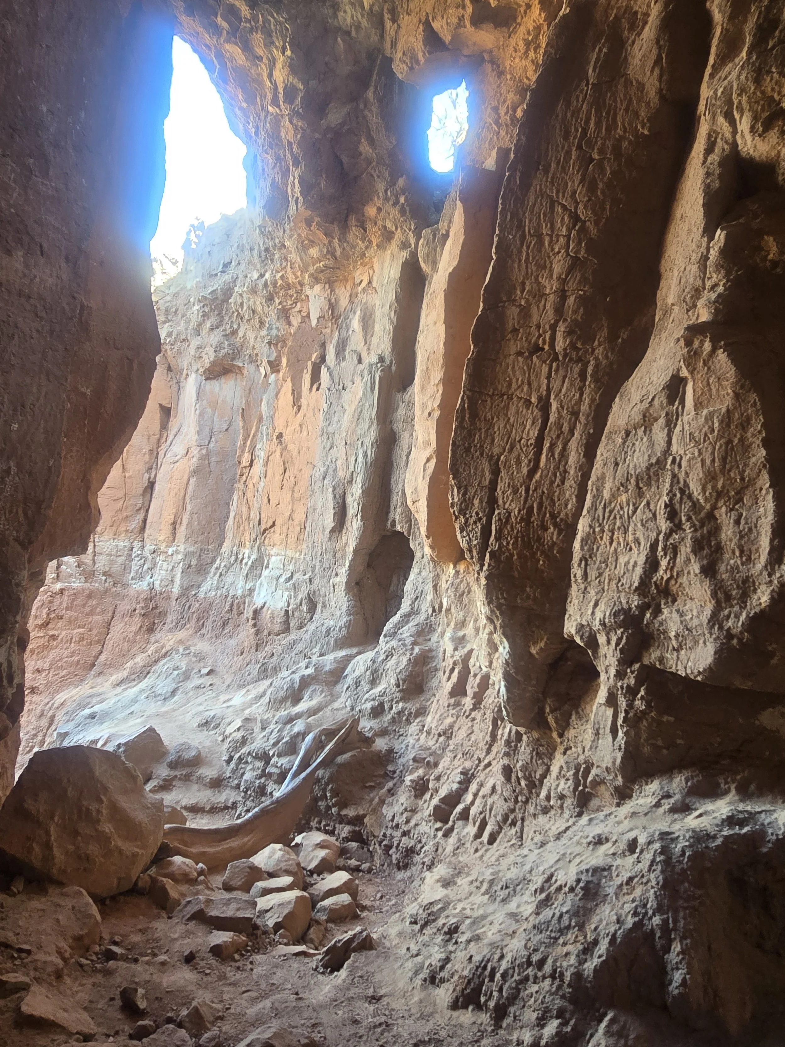 Inside a rocky canyon or cave with sunlight streaming in from the openings at the top, showing rugged reddish-brown rock walls, loose stones, and dust on the ground.