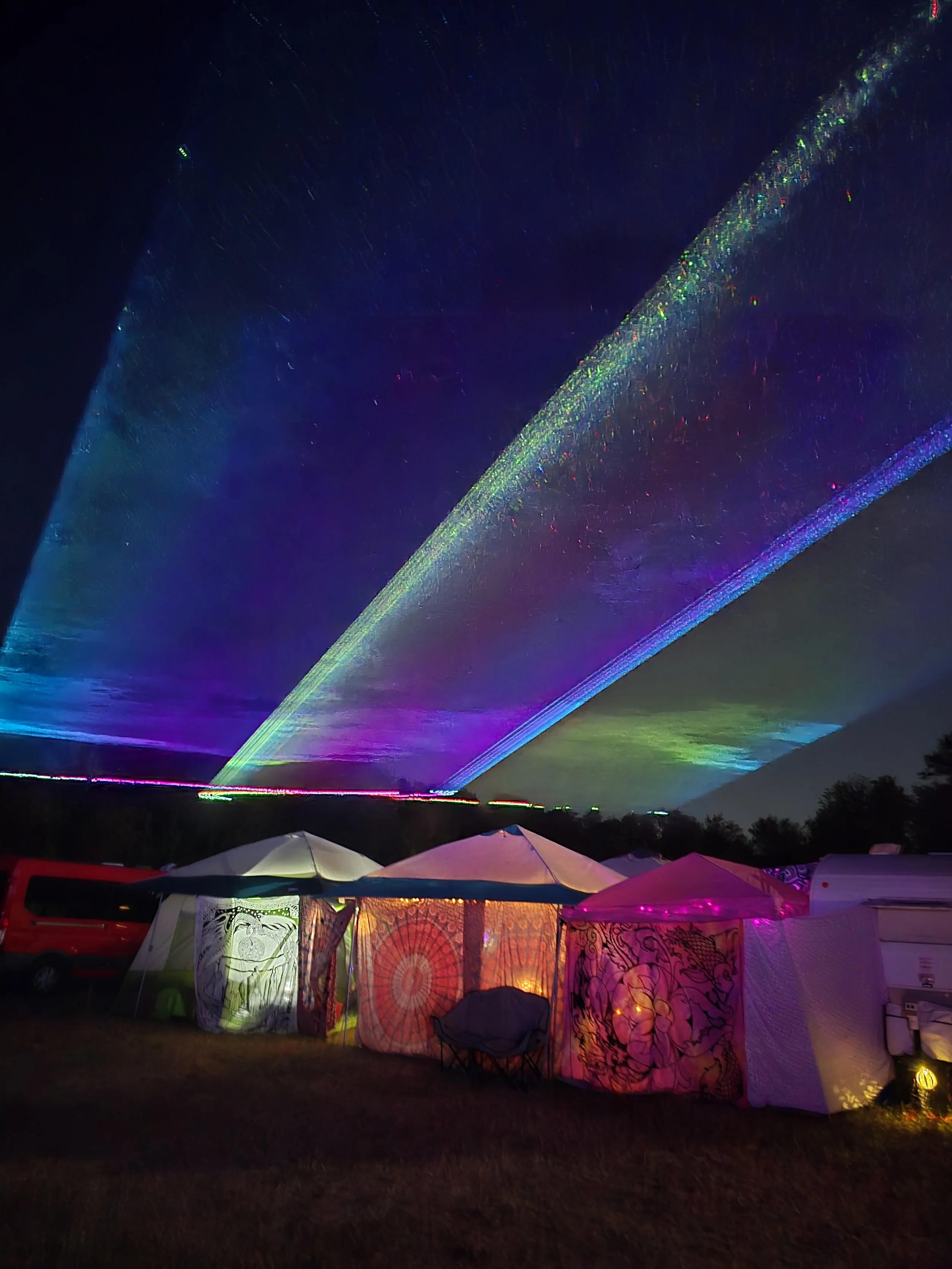 Night scene with colorful laser lights in the sky above tents at a camping site.