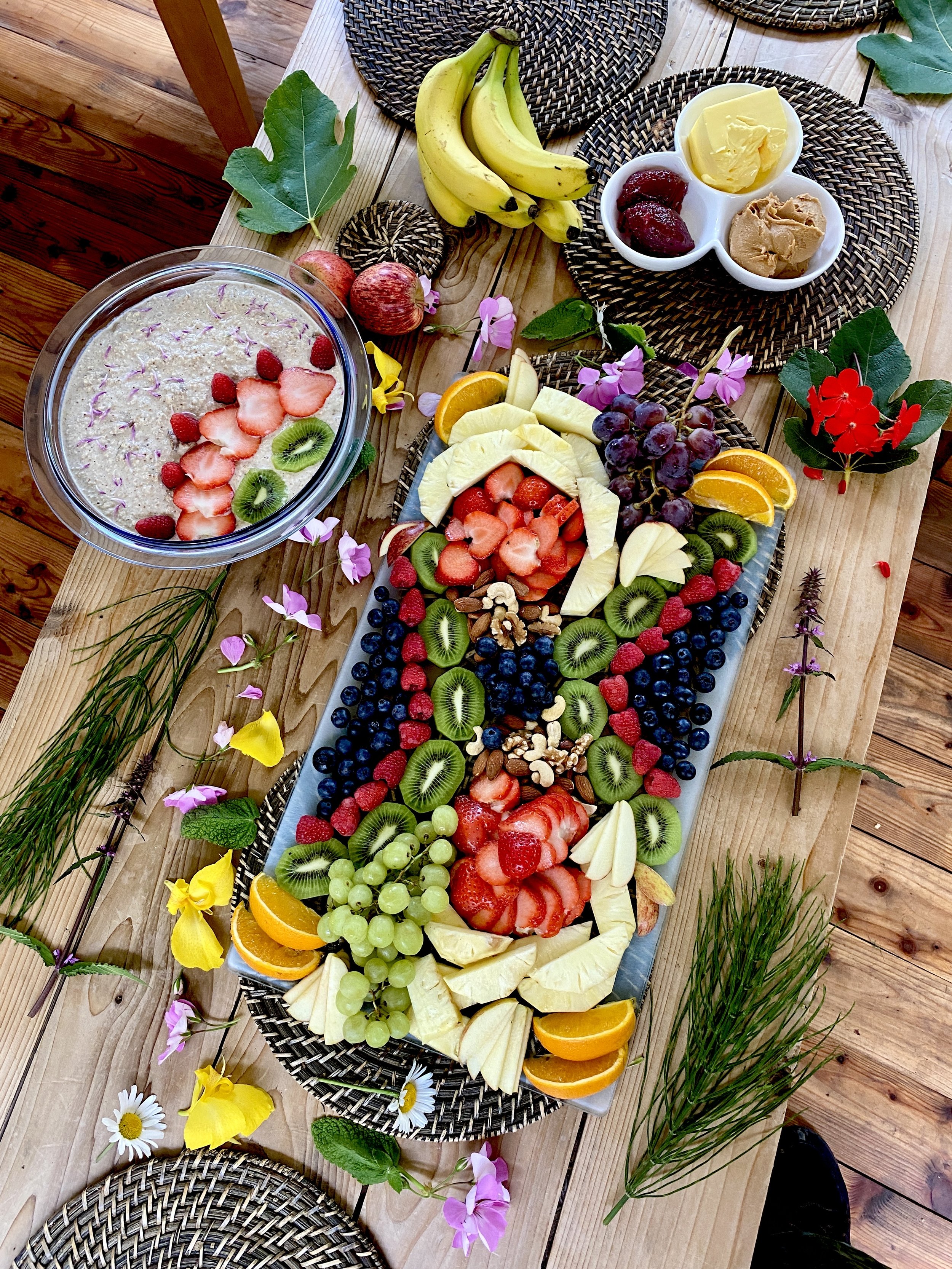 A large serving platter of assorted fresh sliced fruits, including strawberries, kiwi, pineapple, oranges, grapes, and blueberries, arranged in a colorful pattern. Surrounding the platter are bowls of butter, jam, and peanut butter, along with banana