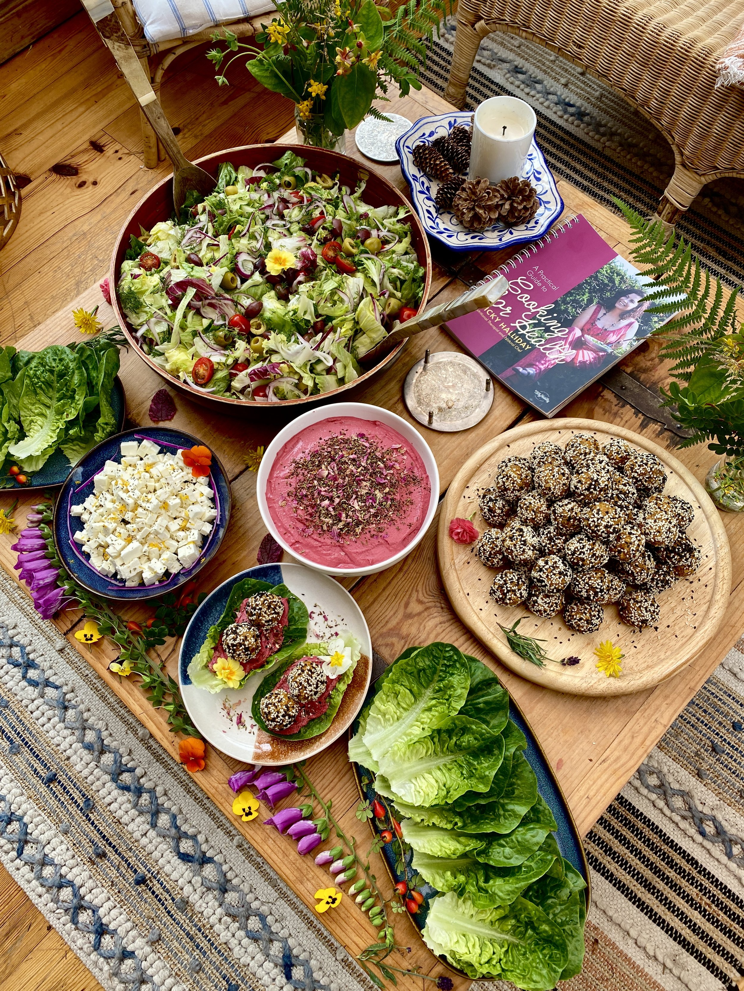 A wooden table set with a colorful salad, a bowl of what appears to be hummus garnished with herbs, a plate of stuffed or coated ball-shaped appetizers, a plate of lettuce-wrapped finger foods, a bowl of feta cheese cubes, and a pink beet or berry di