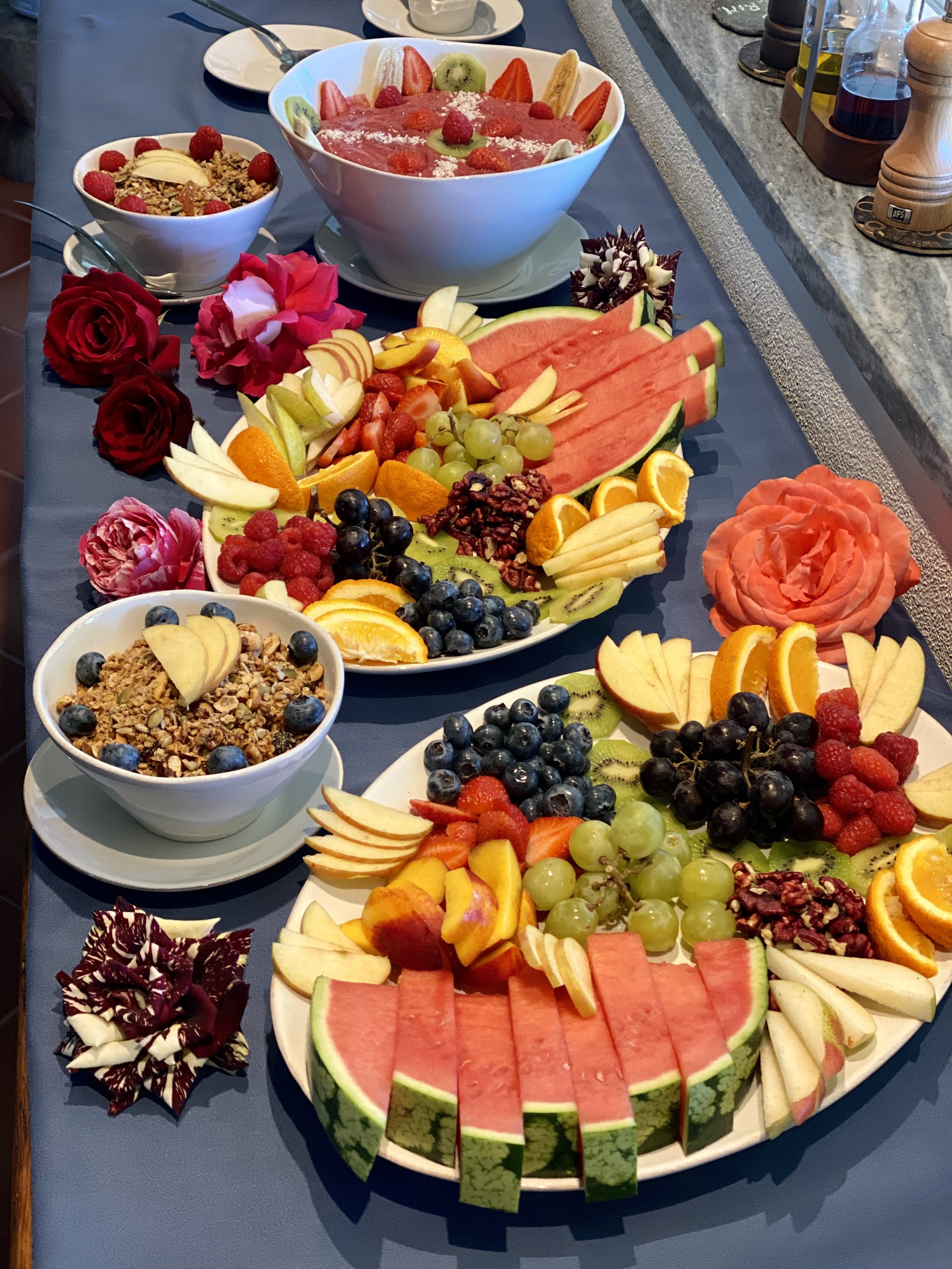 Decorative breakfast table featuring bowls of granola and berries, sliced watermelons and peaches, green and black grapes, strawberries, and flowers.