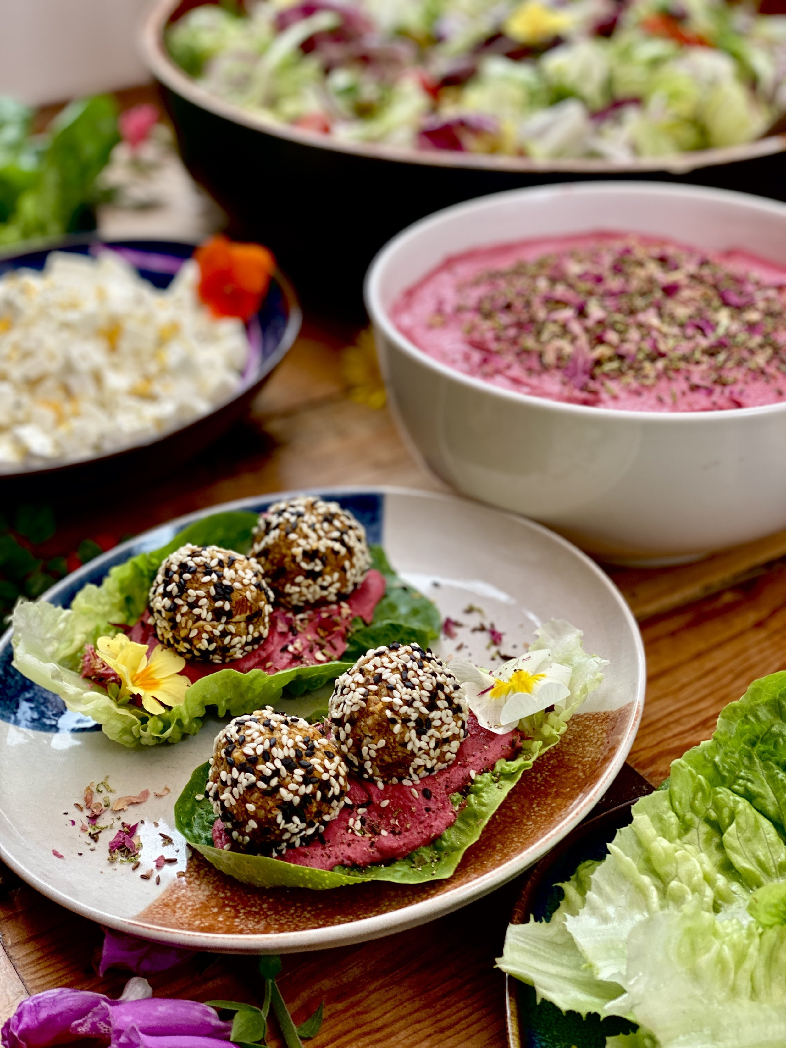 A plate with three falafel balls on lettuce and pink hummus, surrounded by bowls of salad, beet dip, and other appetizers on a wooden table.