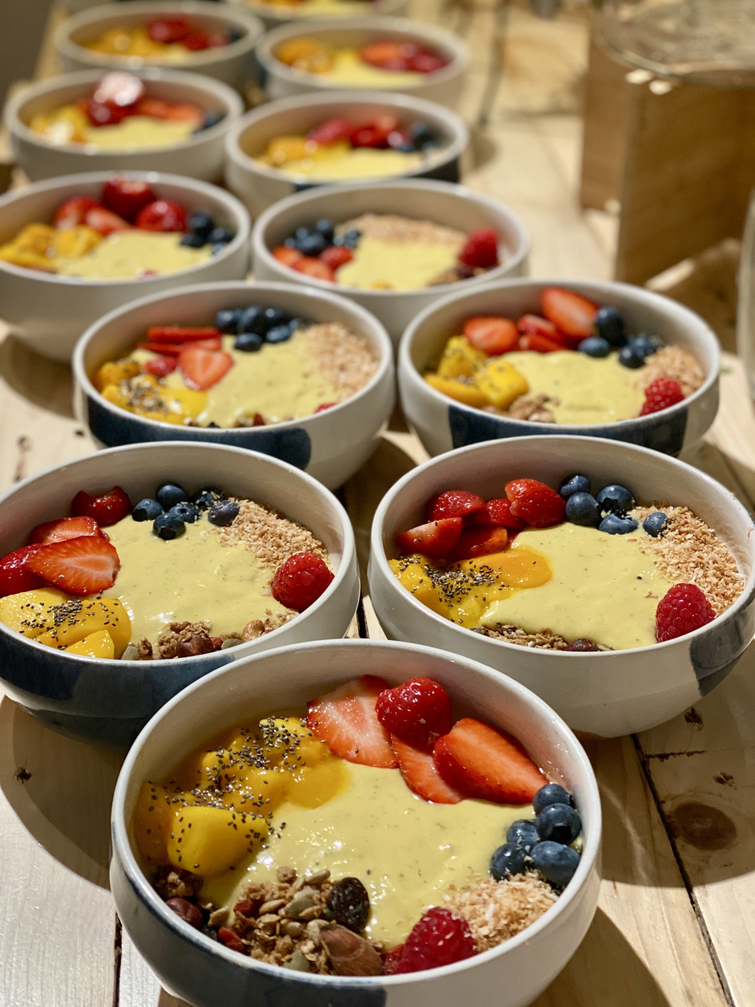 Several bowls of smoothie bowls topped with sliced strawberries, blueberries, raspberries, chopped nuts, seeds, and possibly granola, arranged on a wooden table.