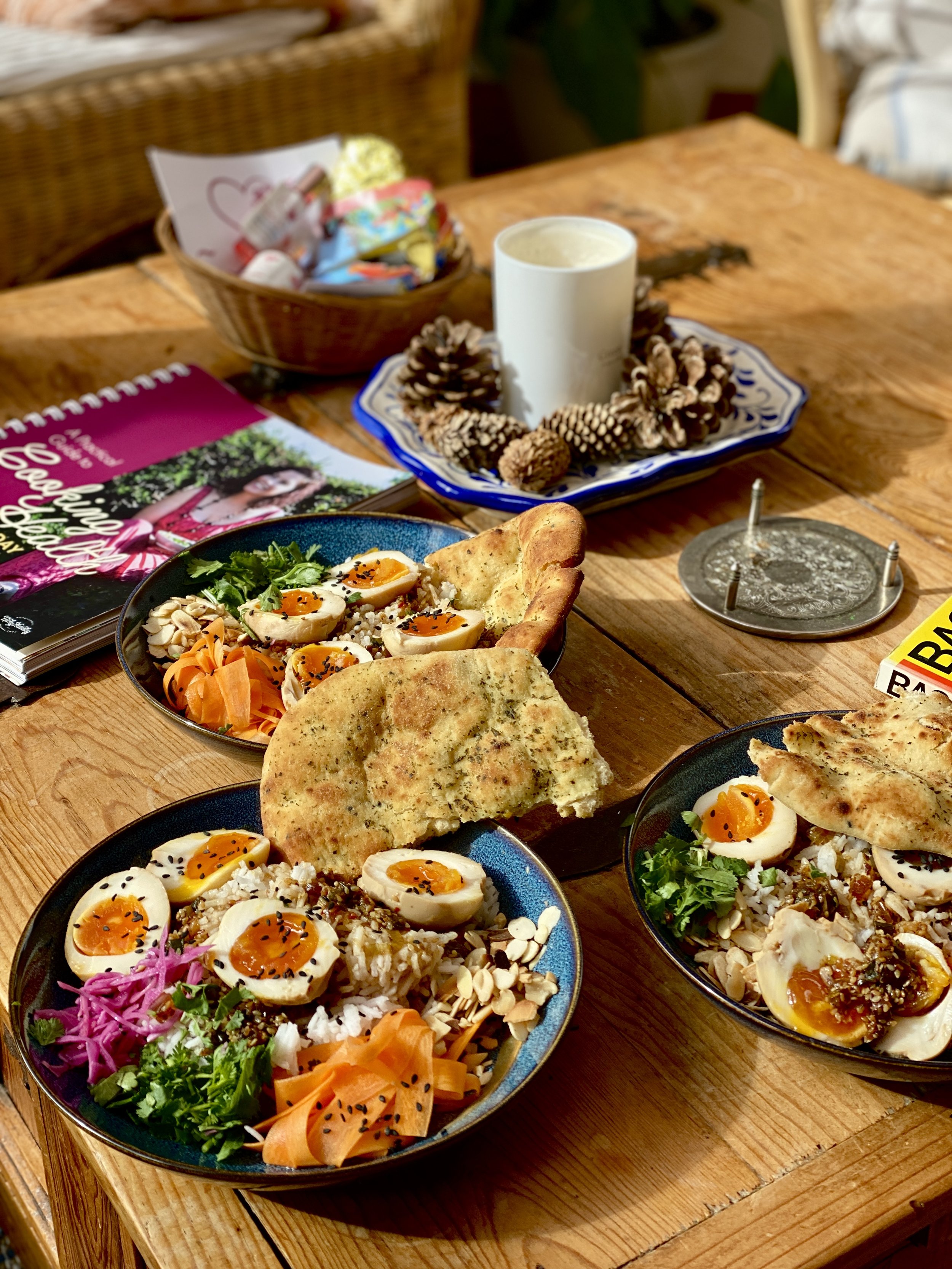 Three bowls of salads with boiled eggs, noodles, vegetables, and flatbread on a wooden table, along with a candle decorated with pinecones, a magazine, a basket with small bottles, and a piece of paper.