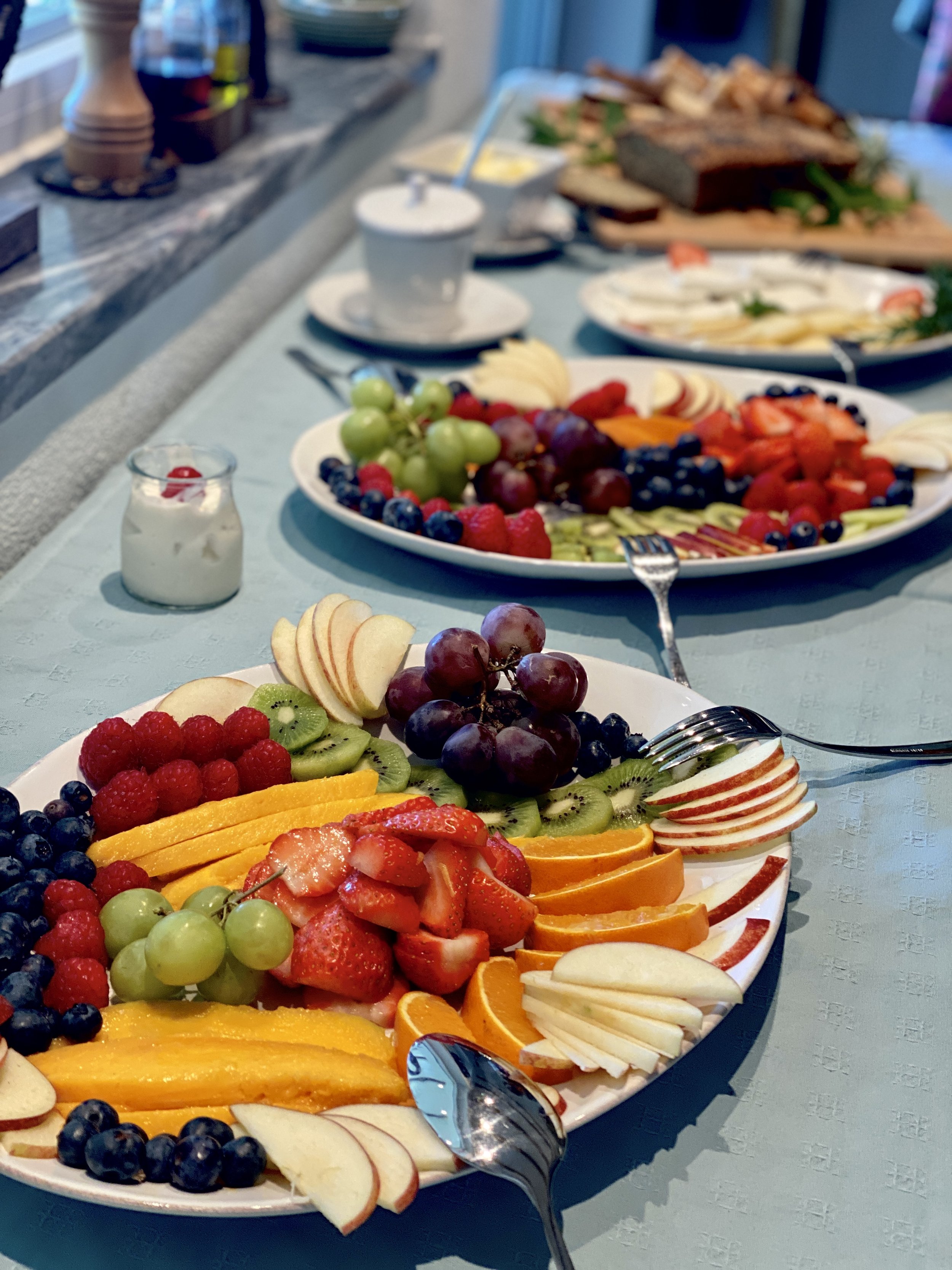 A table set with two large plates of assorted fresh fruit, including strawberries, blueberries, grapes, kiwi, oranges, and apples, with a small jar of yogurt nearby and other dishes in the background.