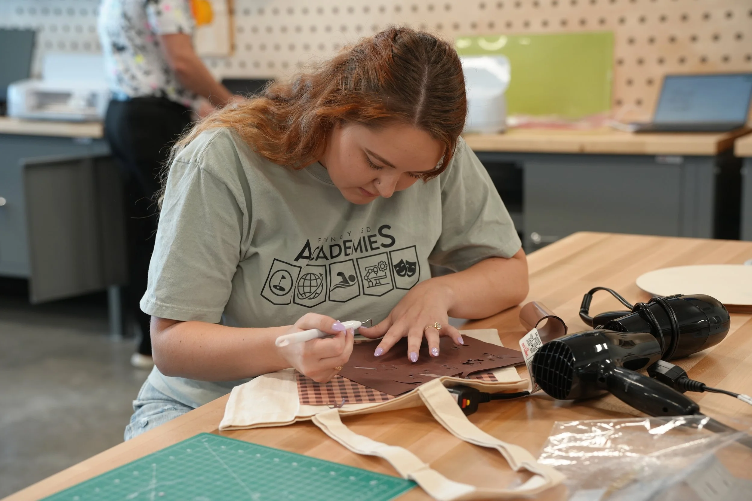 A young woman with red hair is sitting at a wooden table, using a handheld tool to carve or etch a piece of dark brown material. She is wearing a light grey t-shirt with the word "Academies" and various icons printed on it. The table has a green cutting mat, a hairdryer, and other crafting supplies. In the background, another person is standing near a pegboard wall with tools or supplies.