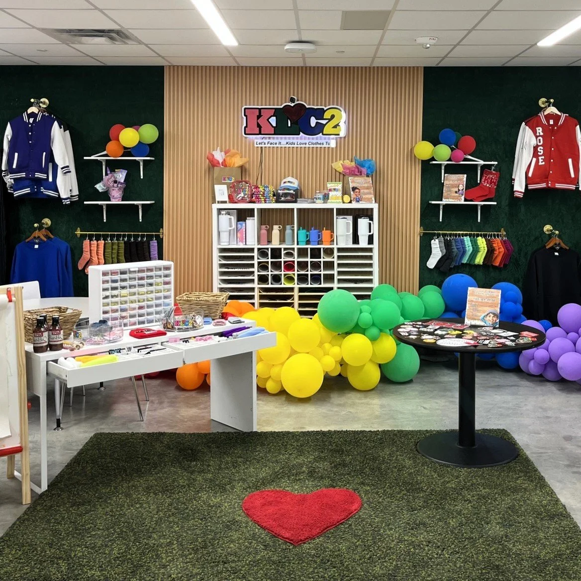 Colorful kids' clothing and accessories display with balloons, jackets, socks, and toys in a store or event setting.