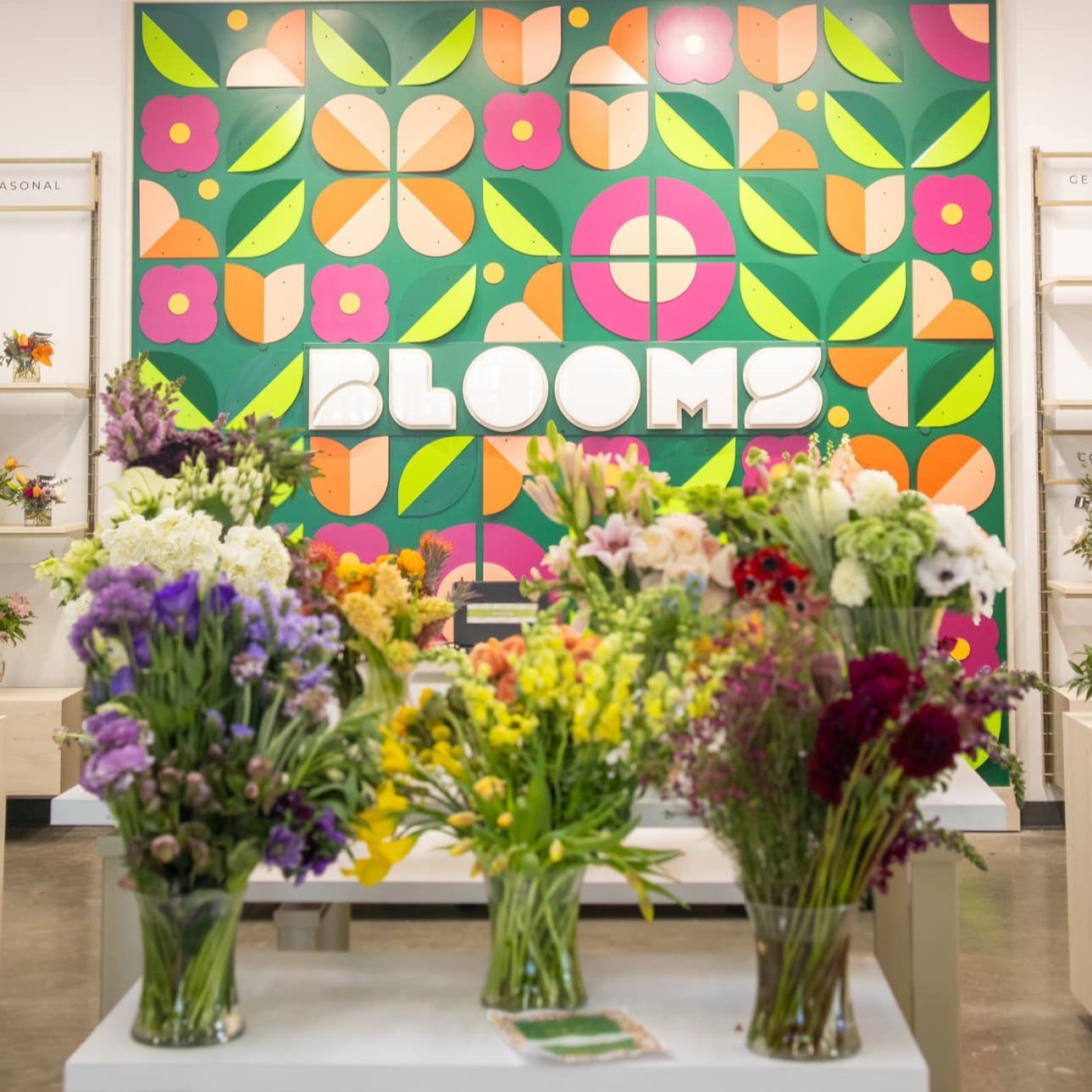 Display of various colorful flowers in vases with a vibrant, patterned floral backdrop spelling out 'BLOOMS' at a flower shop or floral event.
