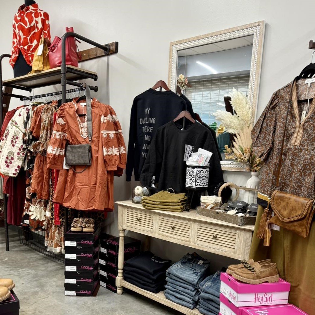 Clothing display in a store with dresses, sweatshirts, folded jeans, shoes, handbags, and decorative elements like a large mirror and floral arrangement.