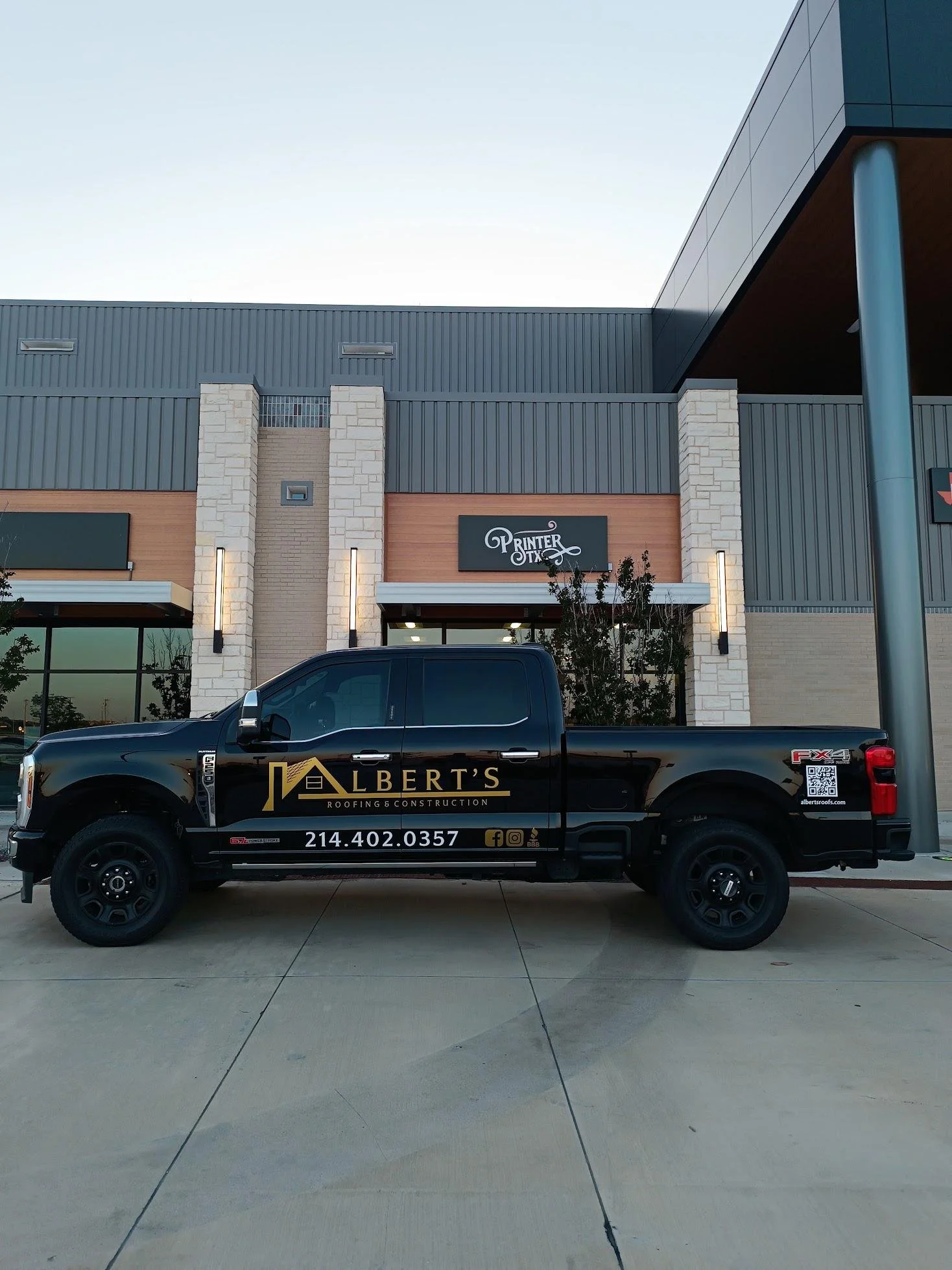 Black pickup truck with advertising for Albert's Roofing & Construction parked in front of a modern commercial building with a sign that reads "Printer TX".