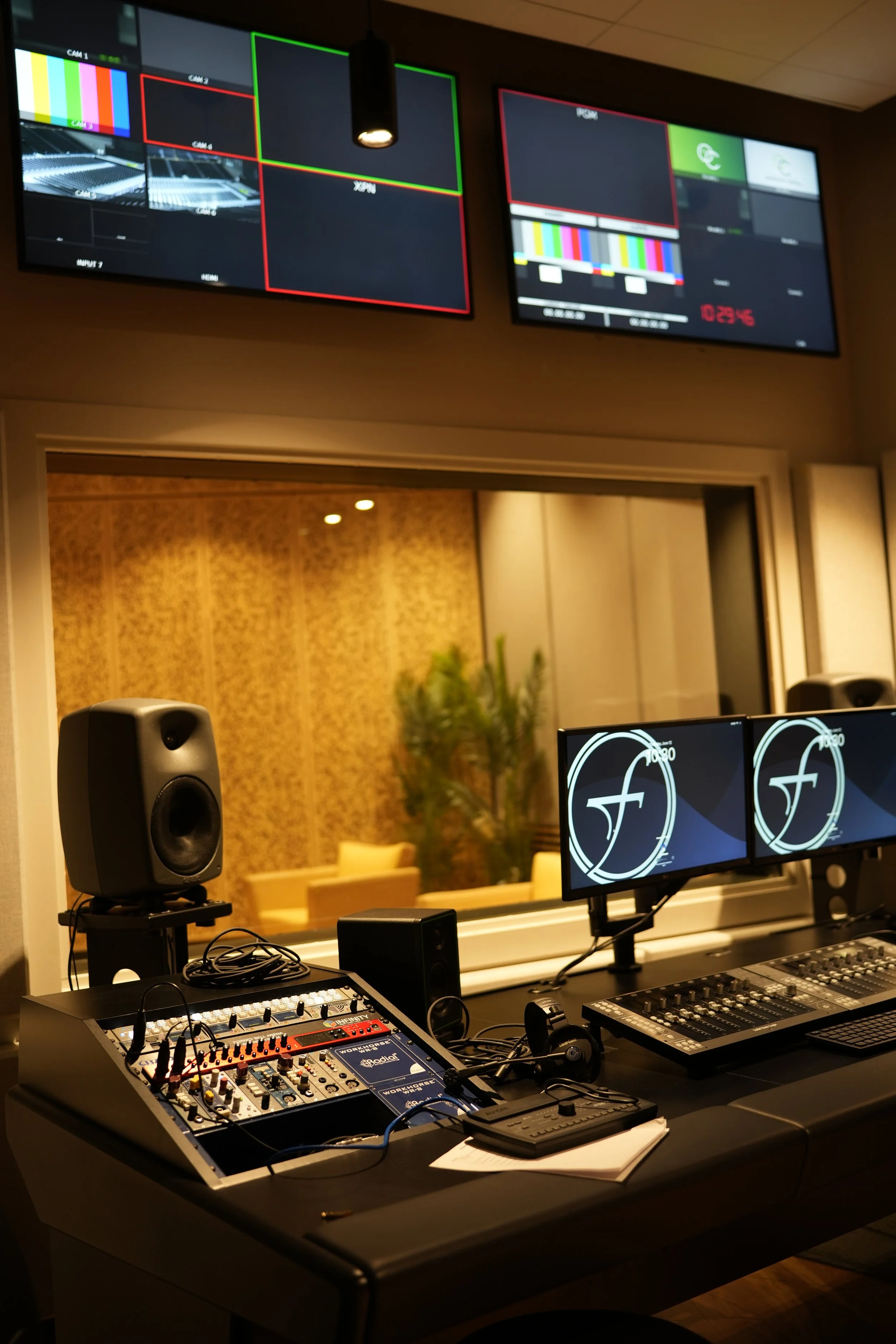 A recording studio control room with mixing consoles, monitors, and speakers, visible through a window from a viewing room with purple and gold patterned walls and seating.