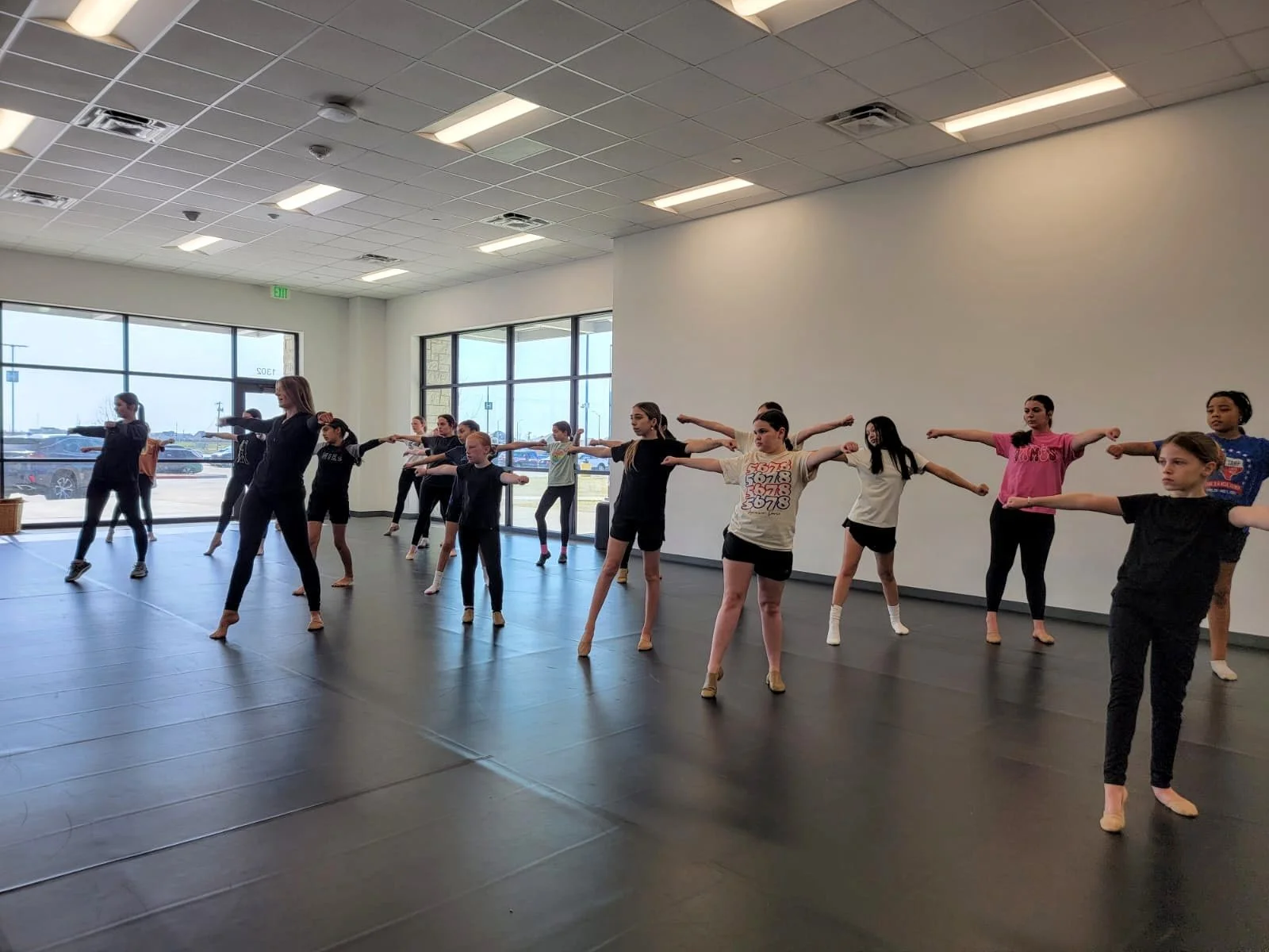A group of young girls practicing ballet in a dance studio with large windows and bright overhead lighting.