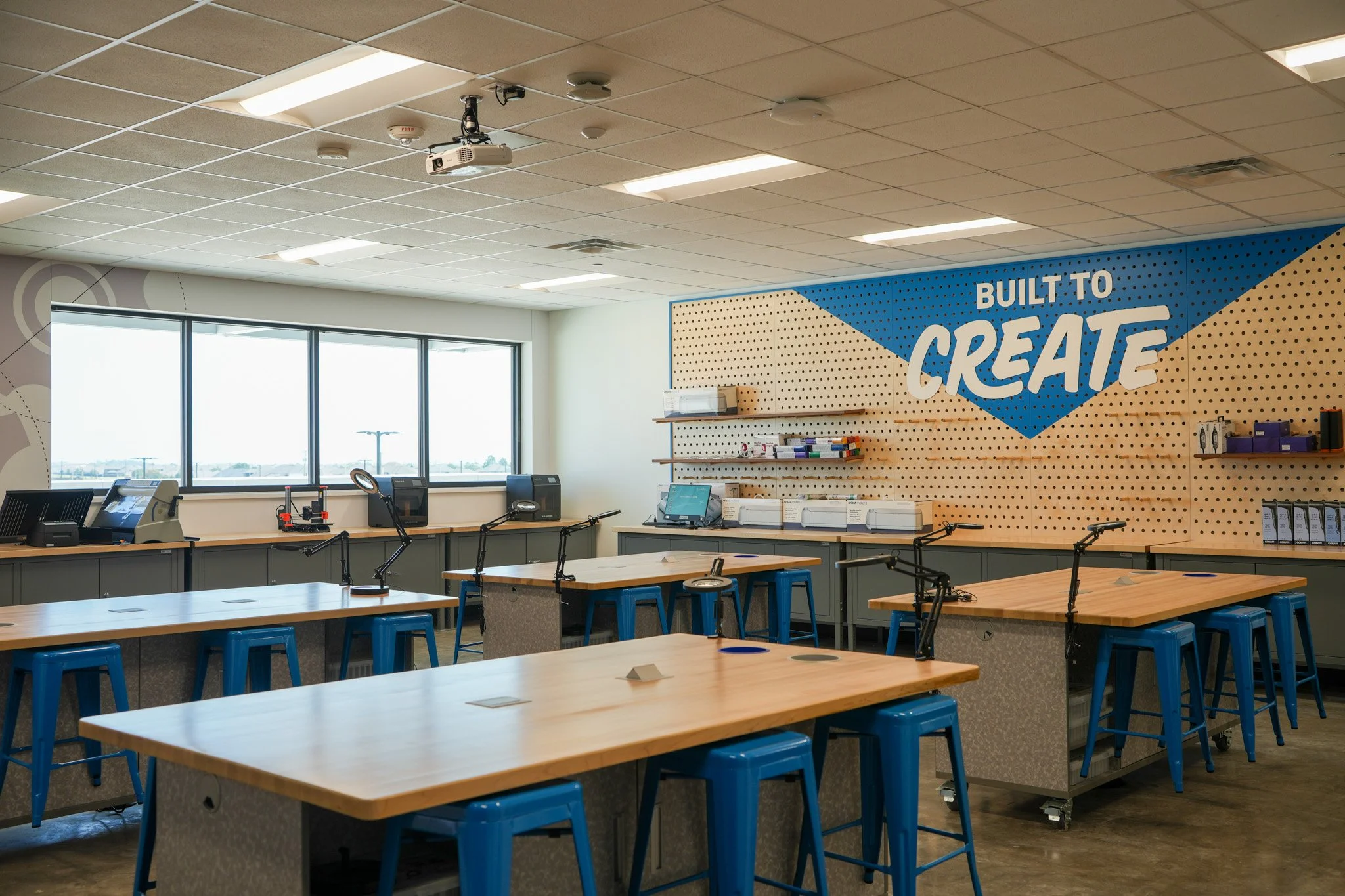 A modern classroom or workshop space with multiple wooden tables and blue stools. The room features a large window, a mounted projector, and a wall with the phrase 'Built to Create' on a blue and beige pegboard.