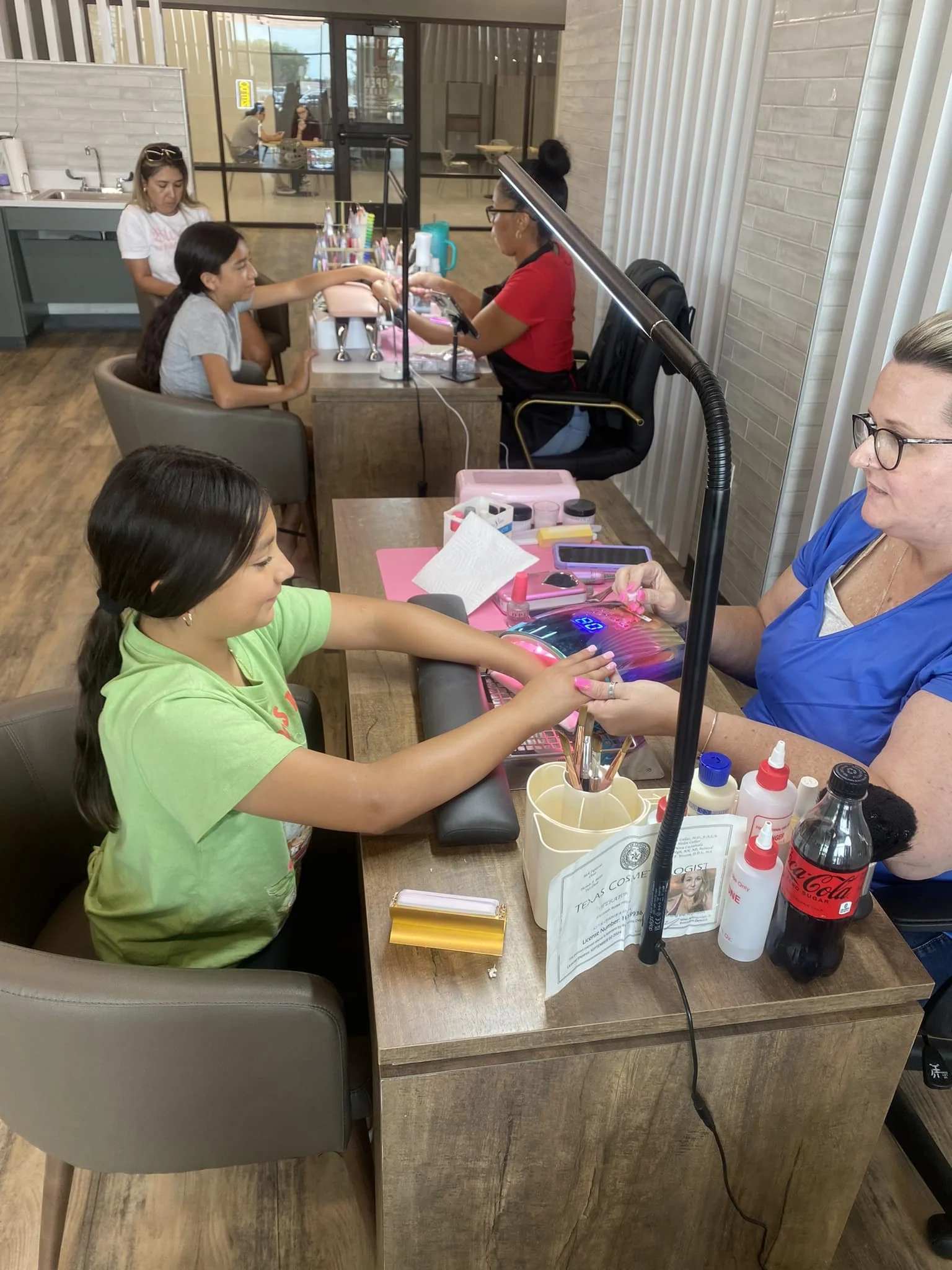 A girl receiving a manicure at a salon with a technician performing the service, while another girl waits in the background.