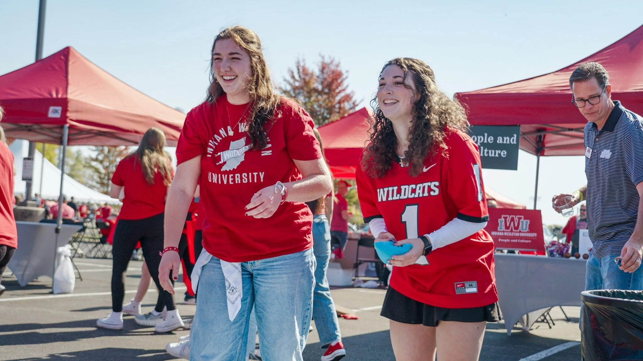 Two young women wearing red Wildcats shirts walking at outdoor event with red tents and people in background.