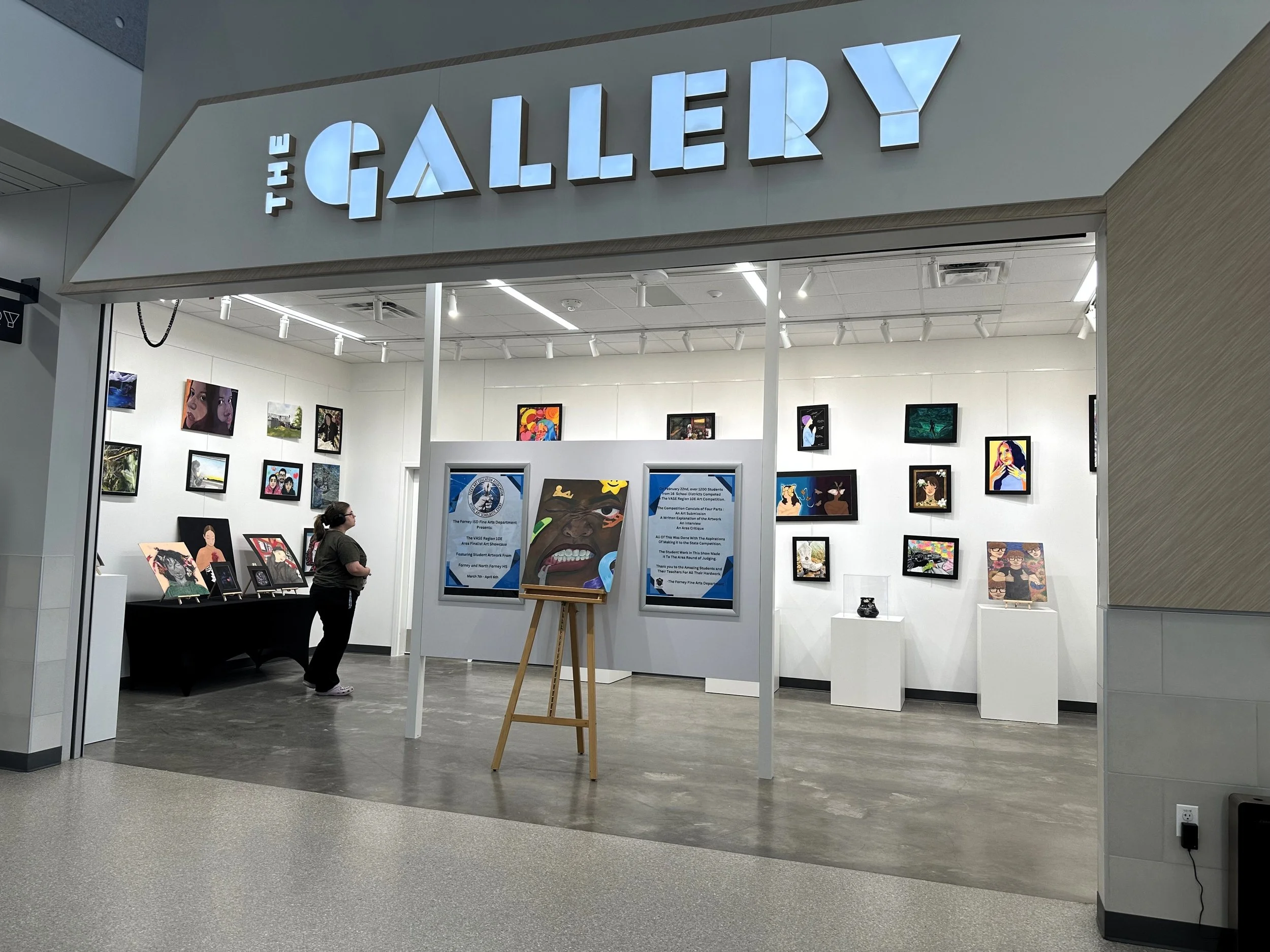View of an art gallery with paintings displayed on white walls and two people observing the artworks.