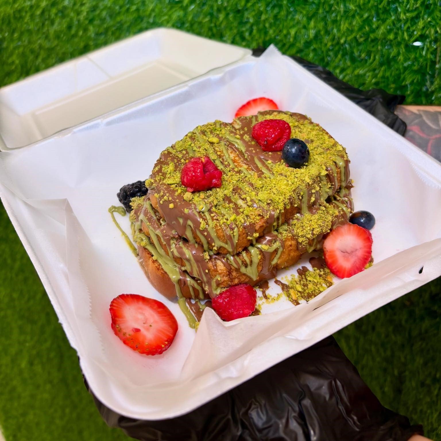 A dessert with fried bread topped with green matcha powder, chocolate drizzle, and fresh berries including strawberries, blueberries, and raspberries, served in a paper-lined box on grass.