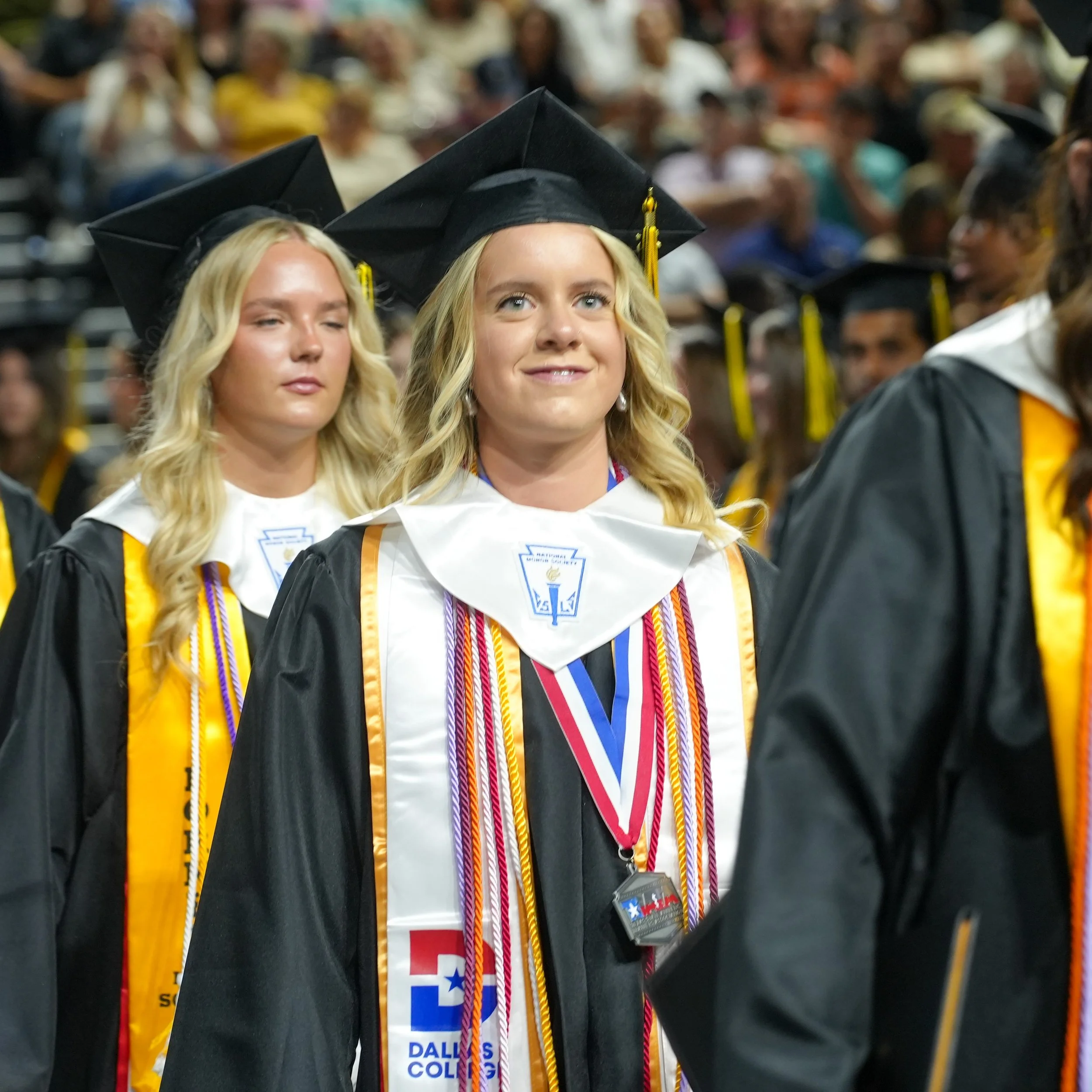 A group of graduates in caps and gowns during a graduation ceremony, with a woman in the foreground wearing a medal and honor cords.