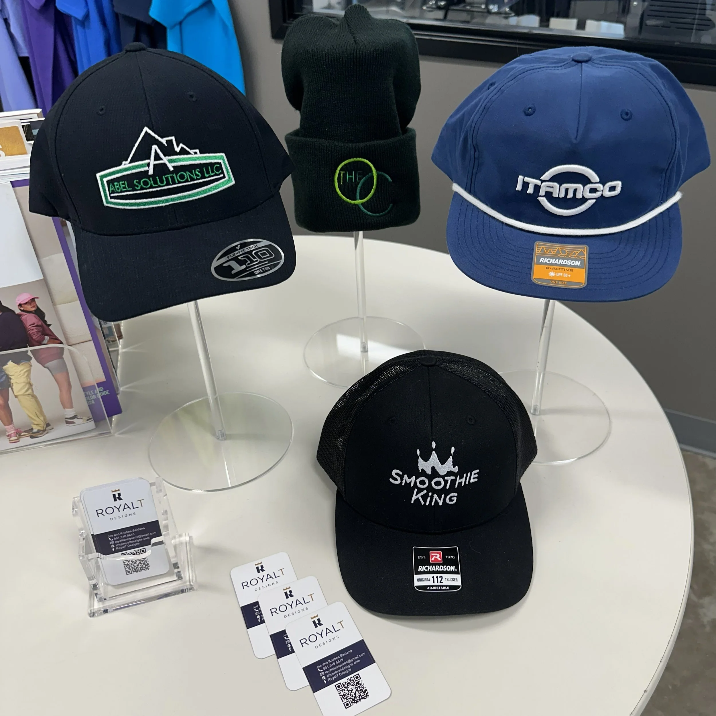 Display of five hats on a white table, including a black trucker hat with a crown logo and 'SMOOTHIE KING' text, three baseball caps with logos for 'ABEL SOLUTIONS LLC', 'ITAMCO', and 'RICHARDSON', and a black knit cap with 'THE' logo.