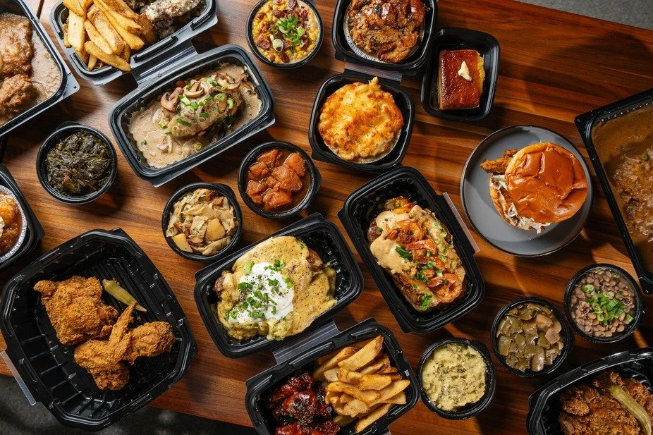 Assorted takeout containers with fried chicken, pasta, mashed potatoes, ribs, burger, and side dishes on a wooden table.