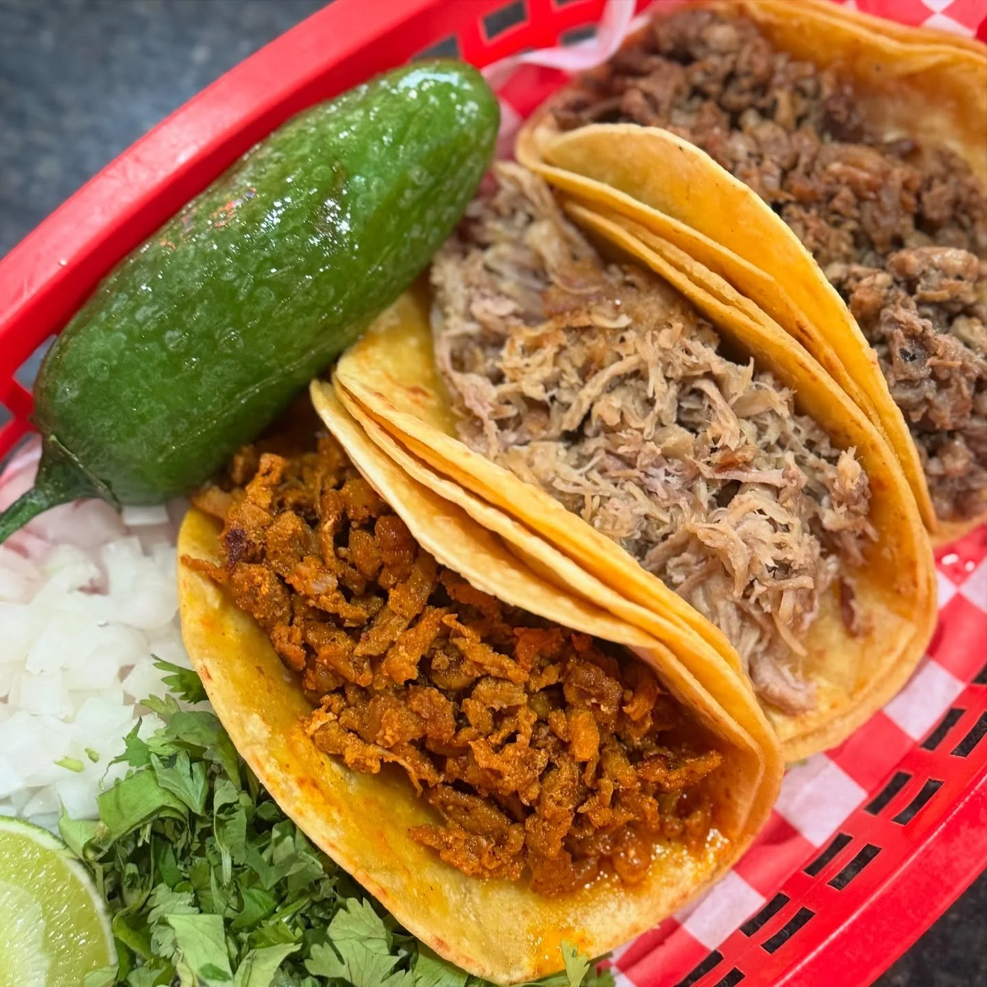 Three tacos filled with shredded meat, ground beef, and chorizo, served with a lime wedge, a jalapeño, chopped onions, and cilantro on a red checkered basket liner.