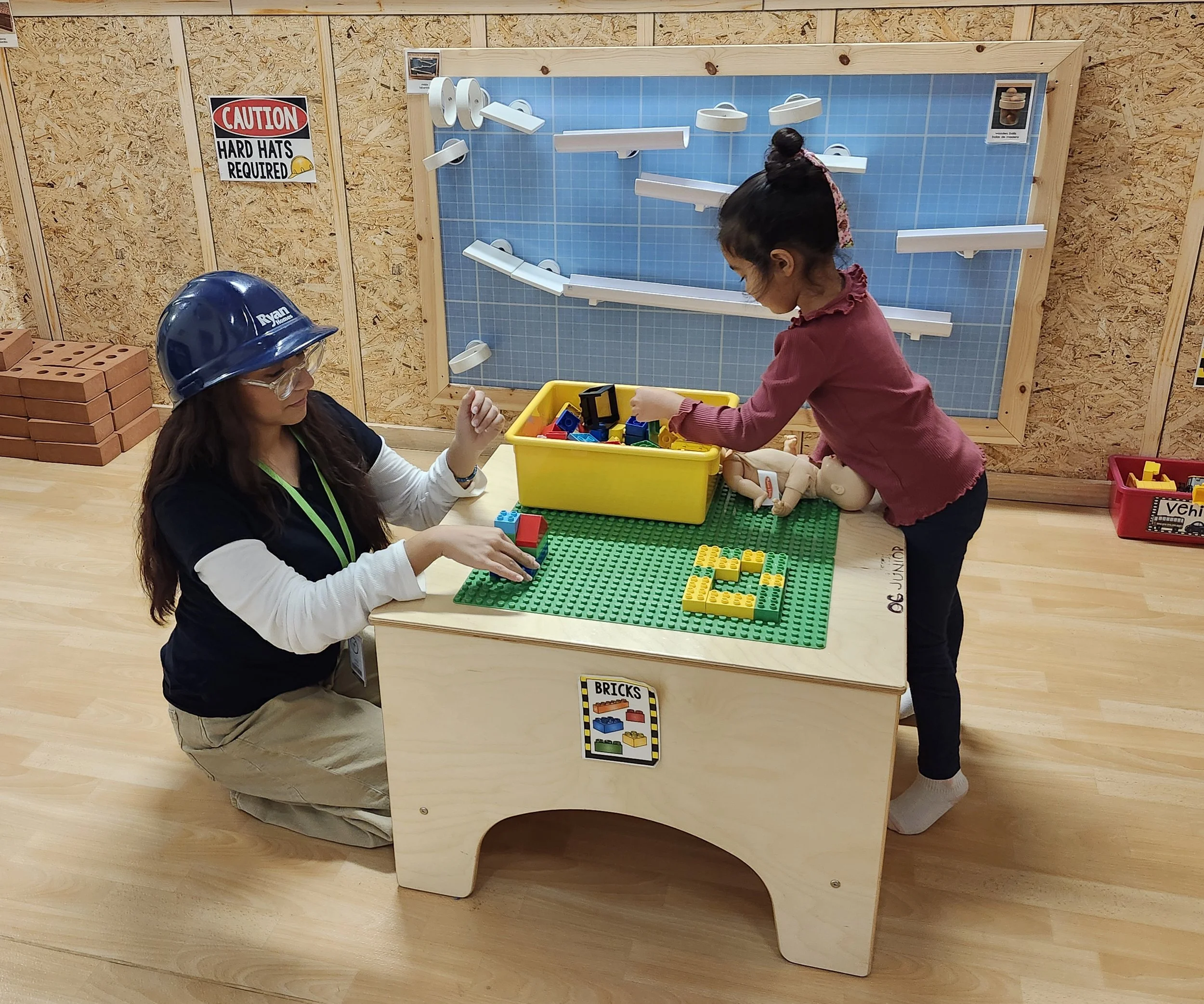 A young girl building with colorful LEGO bricks at a wooden table, supervised by a woman wearing a blue construction helmet and safety glasses in an indoor play area.