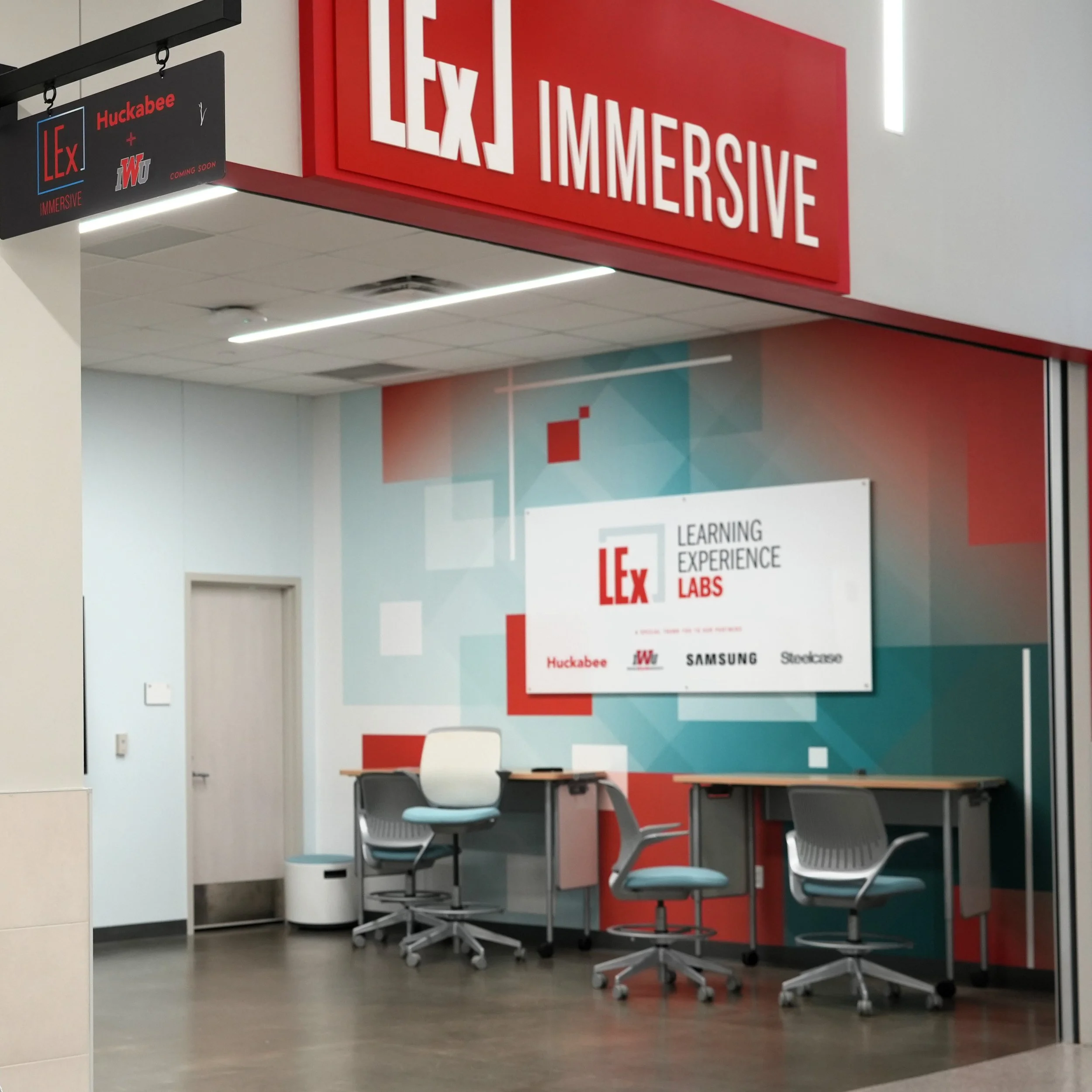 Interior of a learning lab with a wall displaying the logos of Lex Learning Experience Labs, Huckabee, Samsung, and Stokease. There are three chairs, two with wheels, positioned in front of a wall with colorful geometric patterns. A sign and a door are also visible.