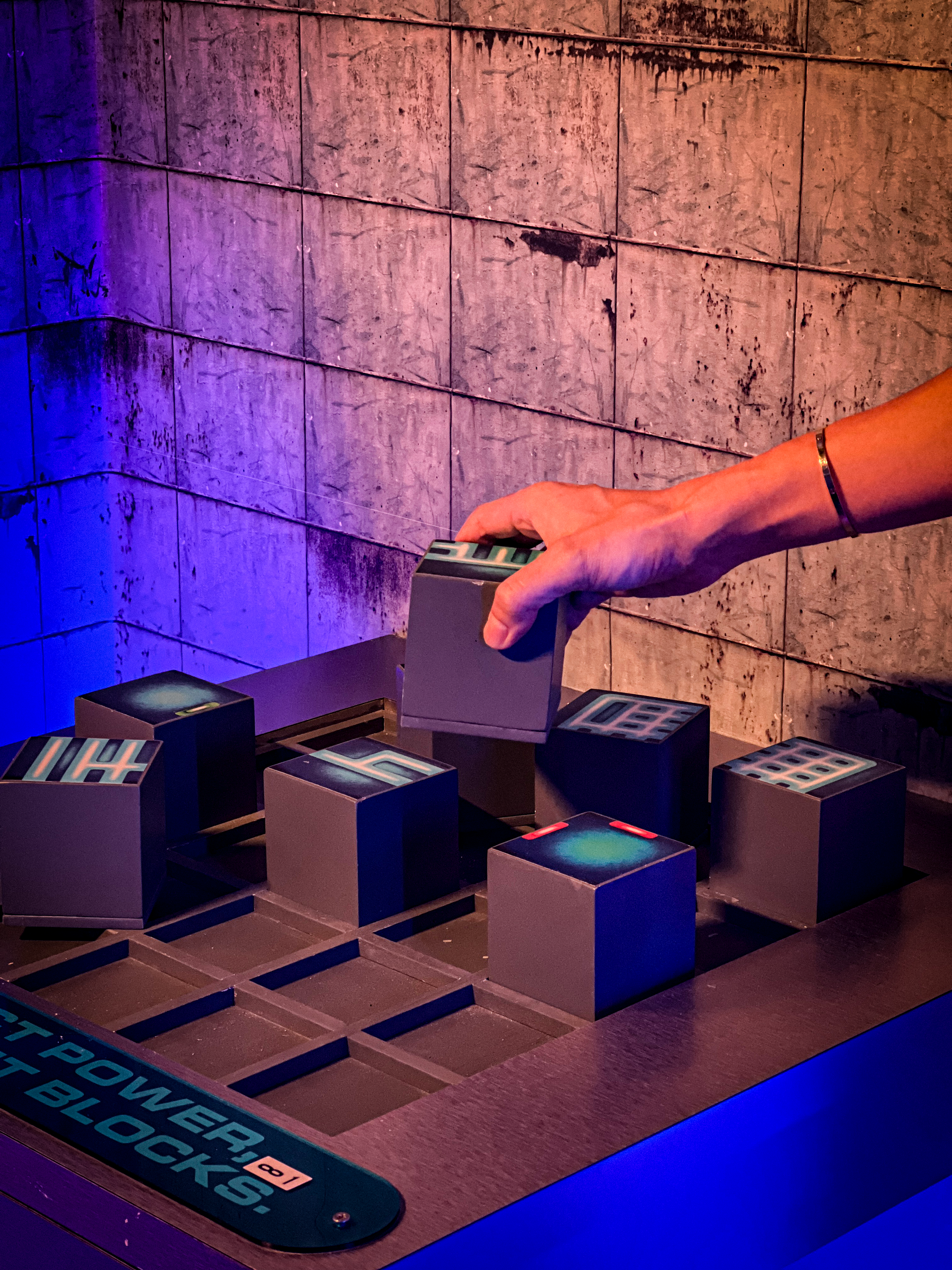 A person's hand is moving a gray block on a black grid in a game or activity with illuminated numbered blocks, colorful lighting, and a textured wall background.