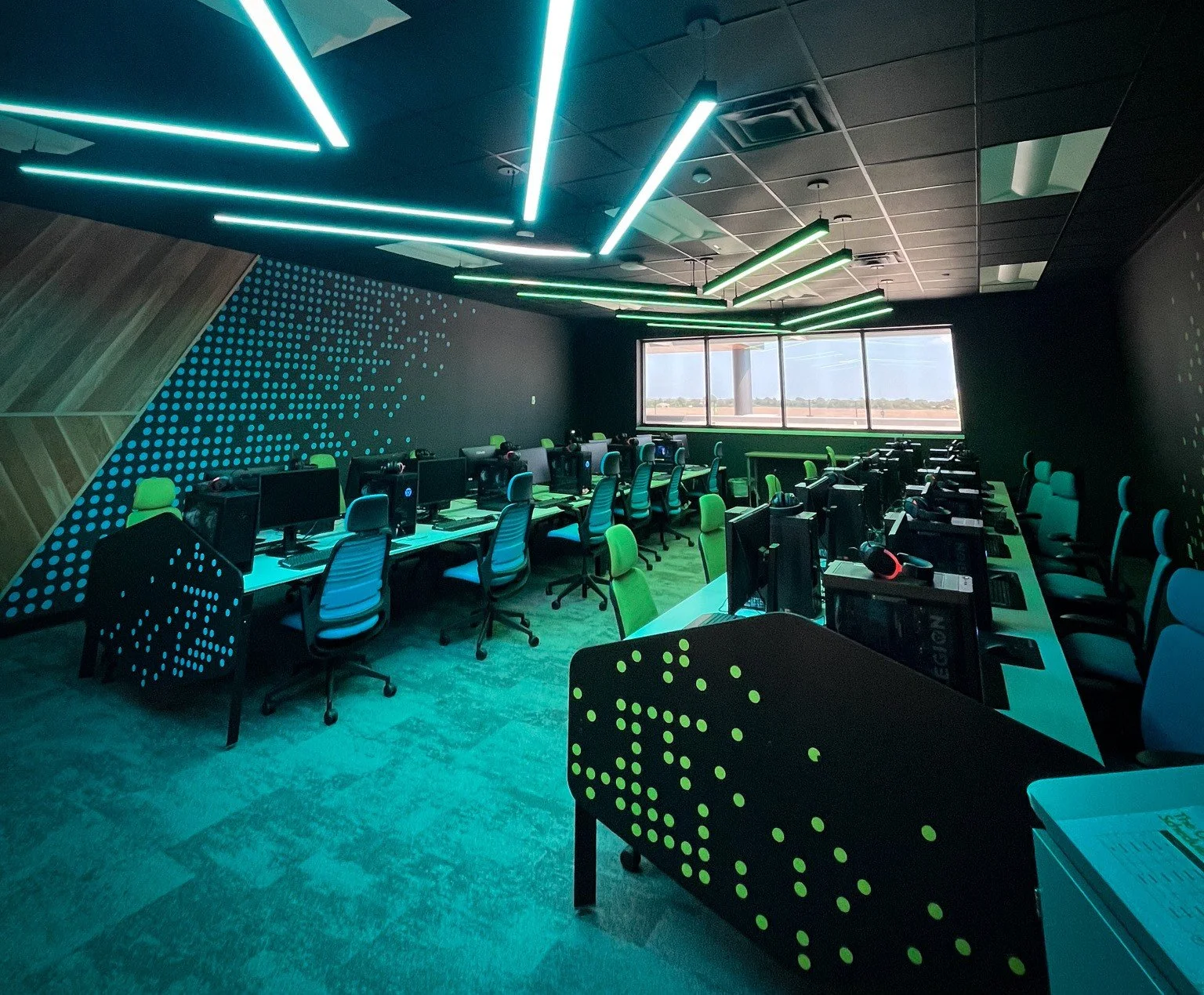 A modern gaming or computer lab with multiple computer station setups, ergonomic chairs, and illuminated green and black decor. Large windows at the far end provide natural light, and ceiling lighting features green and white LED light bars.
