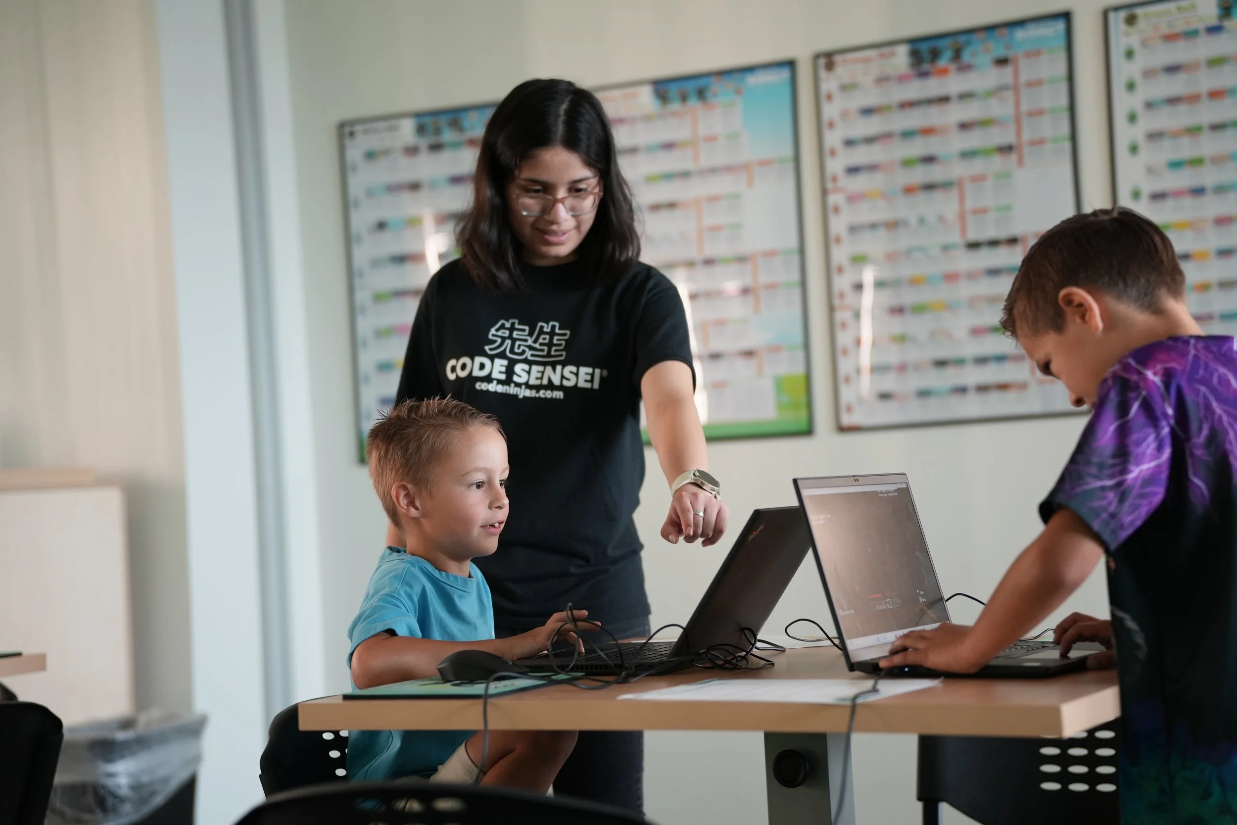 A teacher assisting two boys working on laptops in a classroom with colorful educational posters on the wall.