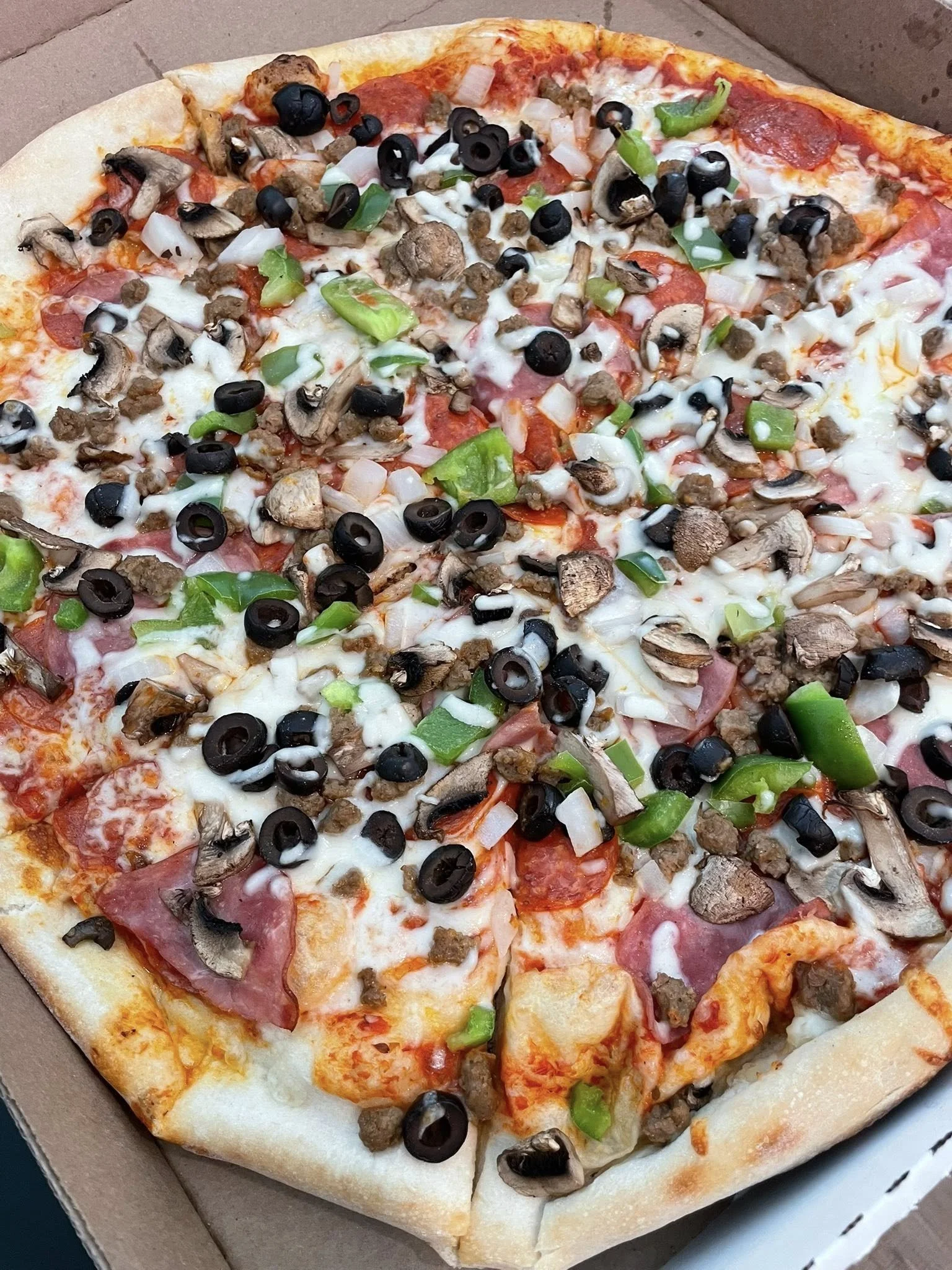 A pepperoni pizza topped with black olives, green bell peppers, mushrooms, onions, and sausage in a cardboard pizza box.