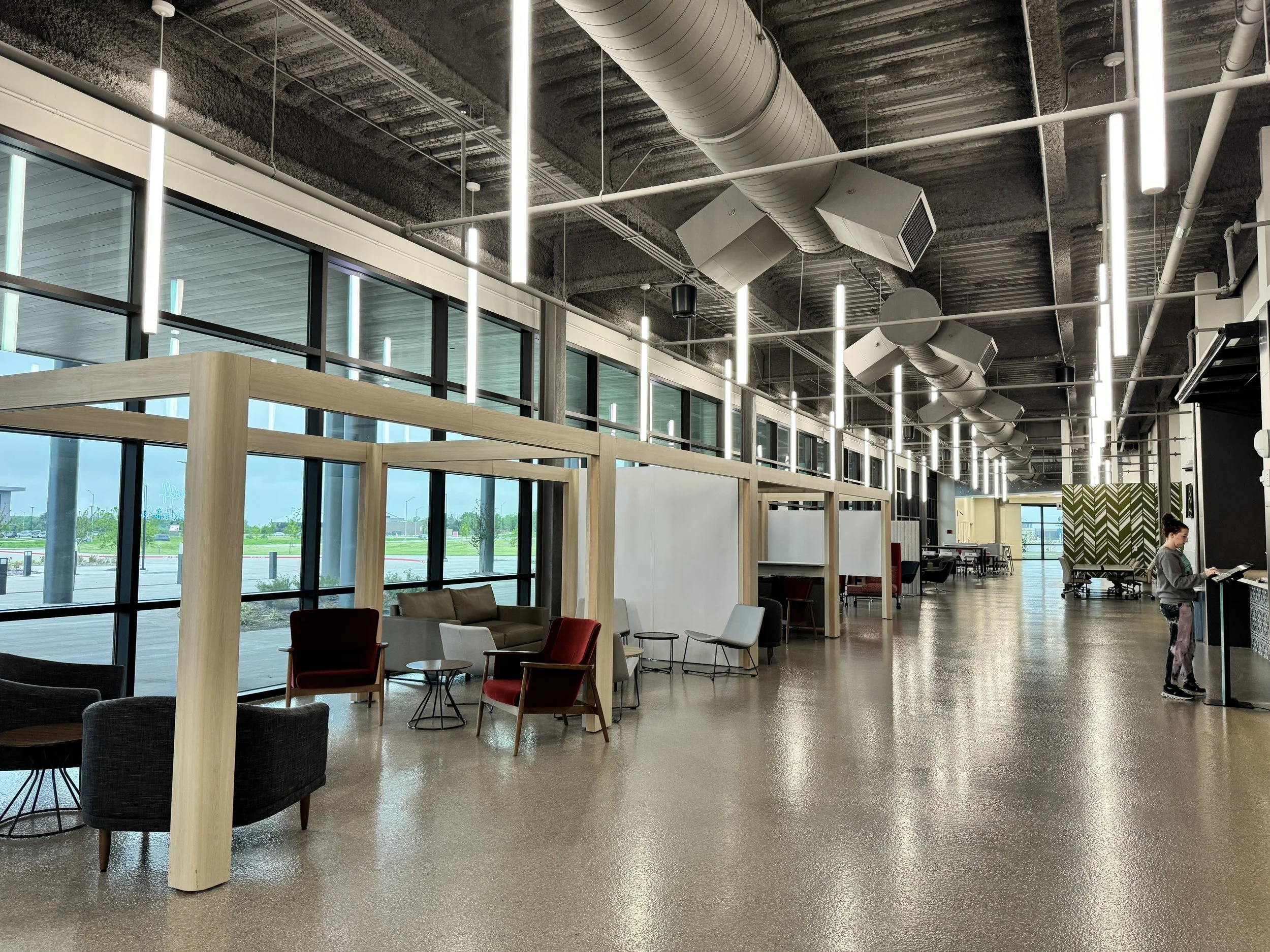 Modern airport waiting lounge with seating areas, large windows, and high ceiling with exposed ductwork and lighting.