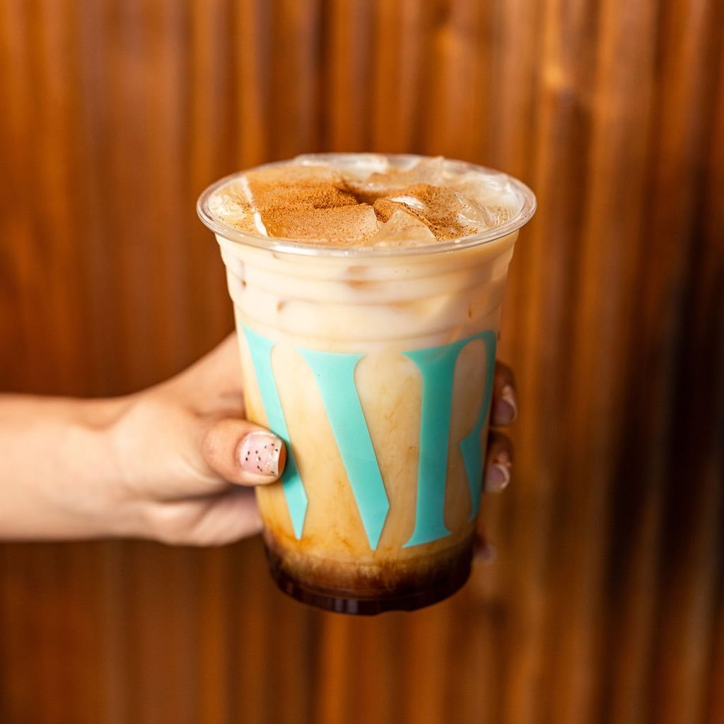 A hand holding a plastic cup filled with iced coffee topped with whipped cream and cinnamon, against a wooden background.