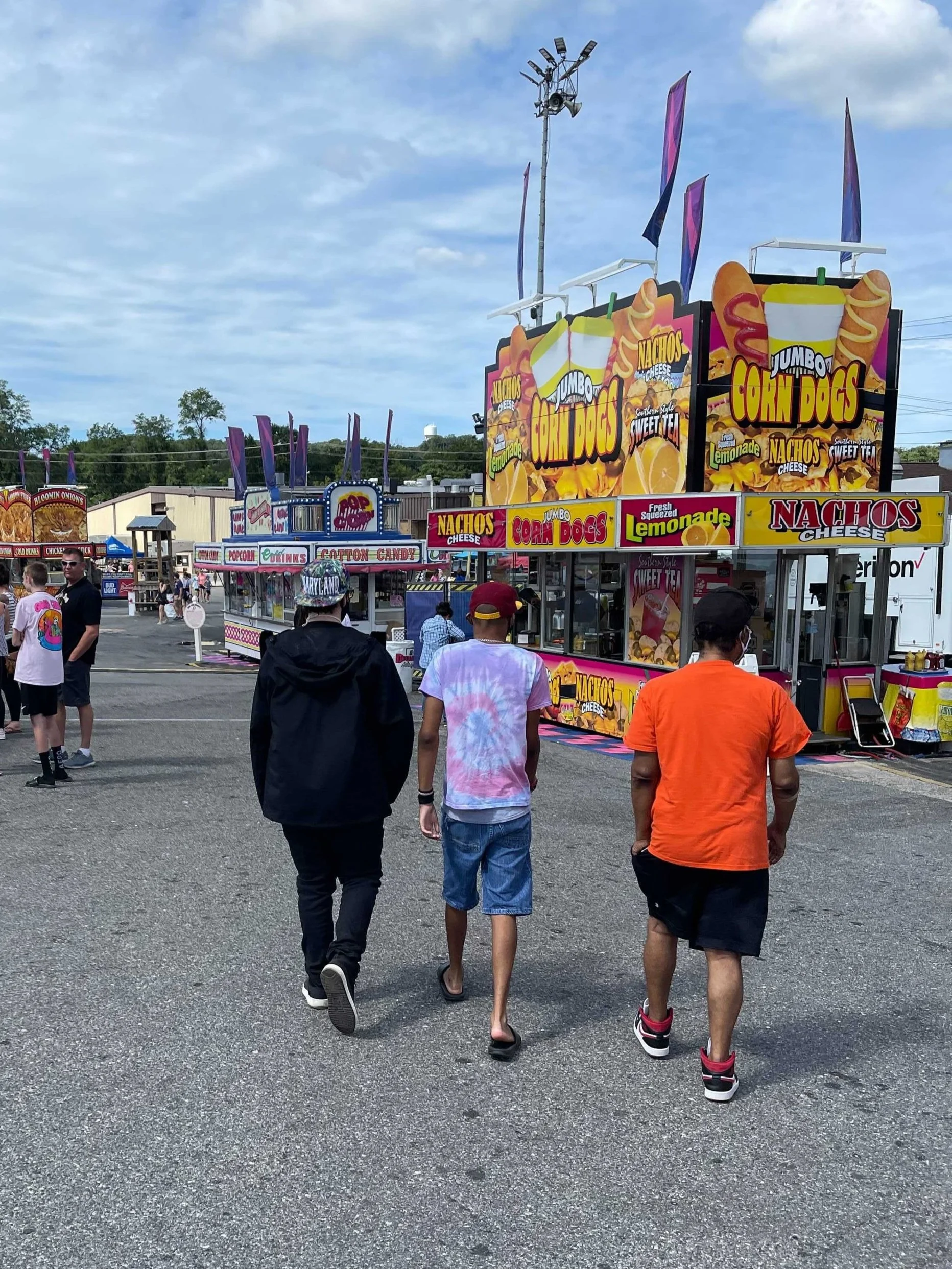 Three men walk around a carnival toward a corn dog stand