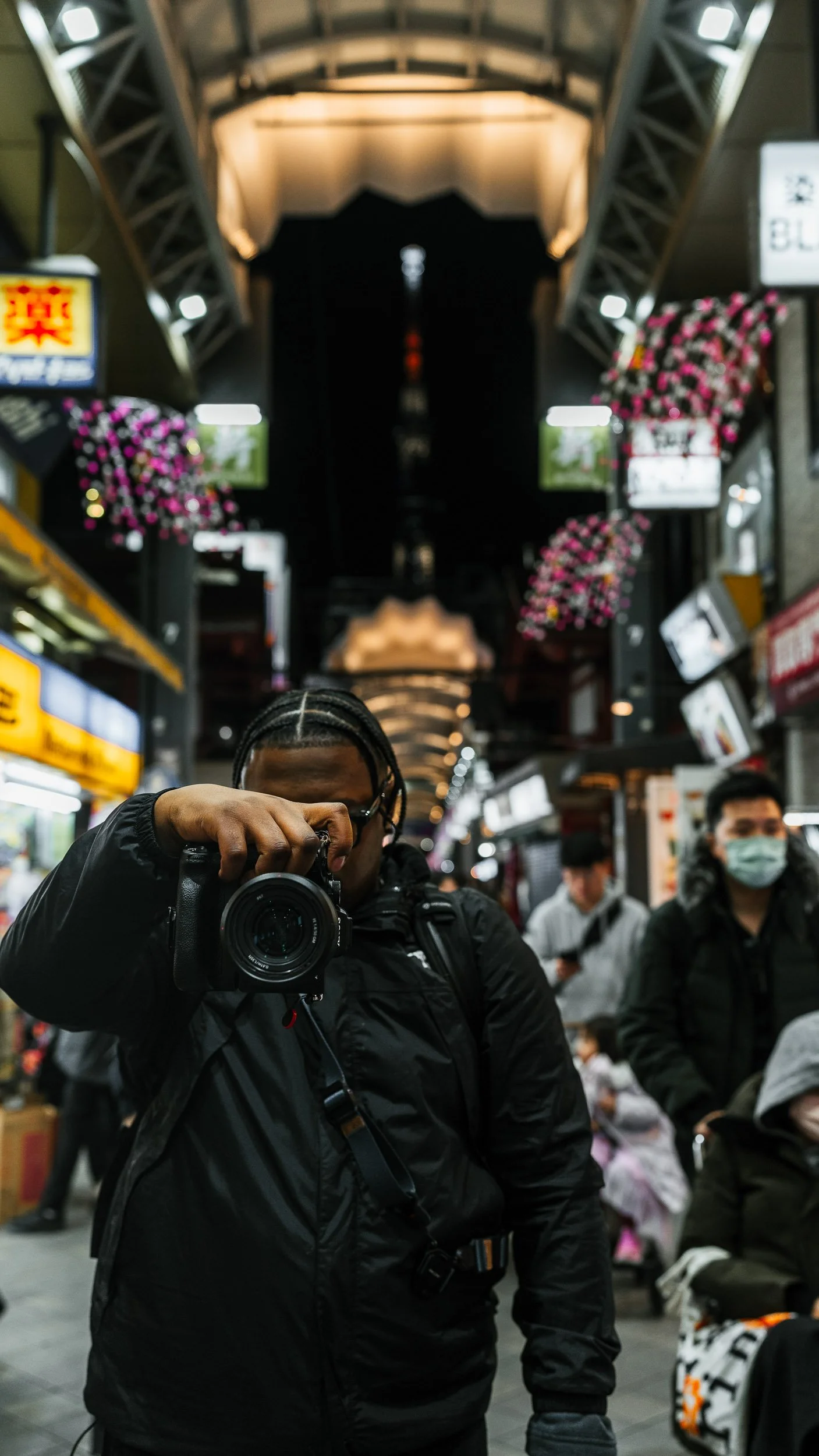 Person with braided hair taking a photo in a busy indoor market or shopping area, surrounded by other people and illuminated signs.
