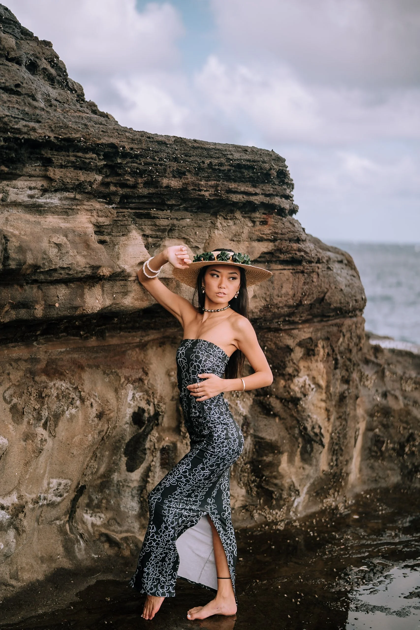 A woman in a strapless dress and wide-brimmed hat stands on a rocky shoreline, with ocean waves in the background, touching her hat with one hand and resting her other hand on her waist.