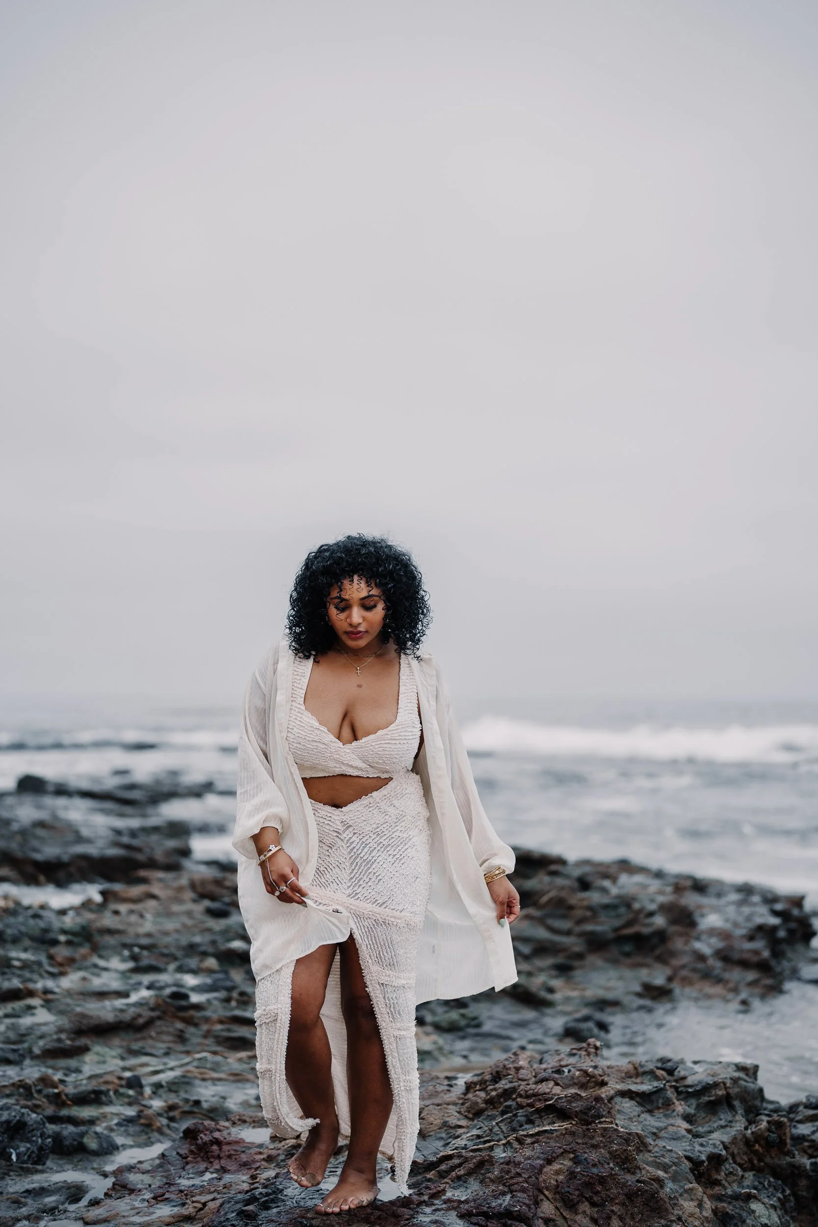 A woman with curly black hair walking barefoot on rocky beach, wearing a white textured dress and a light shawl, with the ocean and overcast sky in the background.