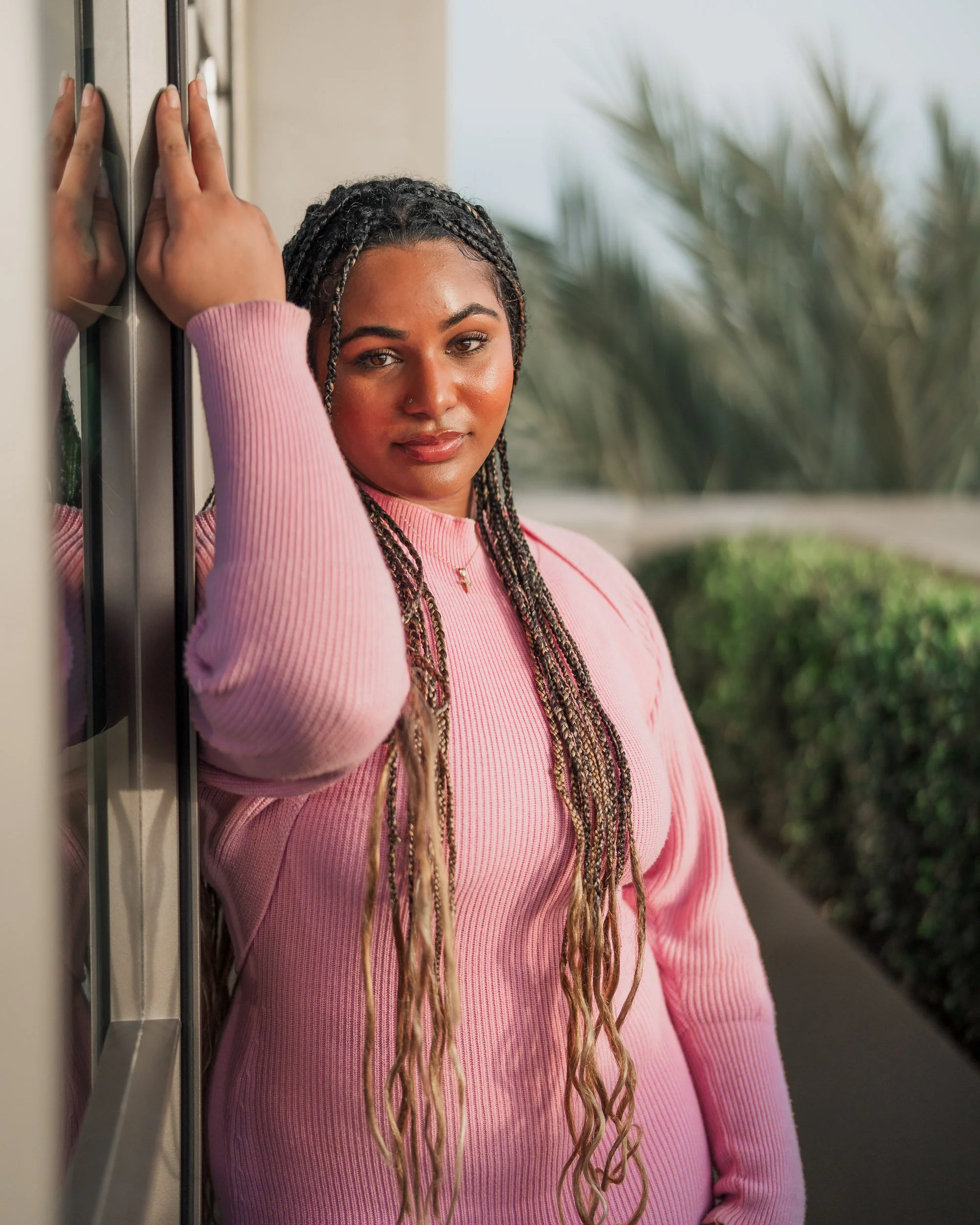 A woman with braided hair and a pink sweater poses with her head resting on her hand next to a glass window. There is greenery and a pathway outside in the background.