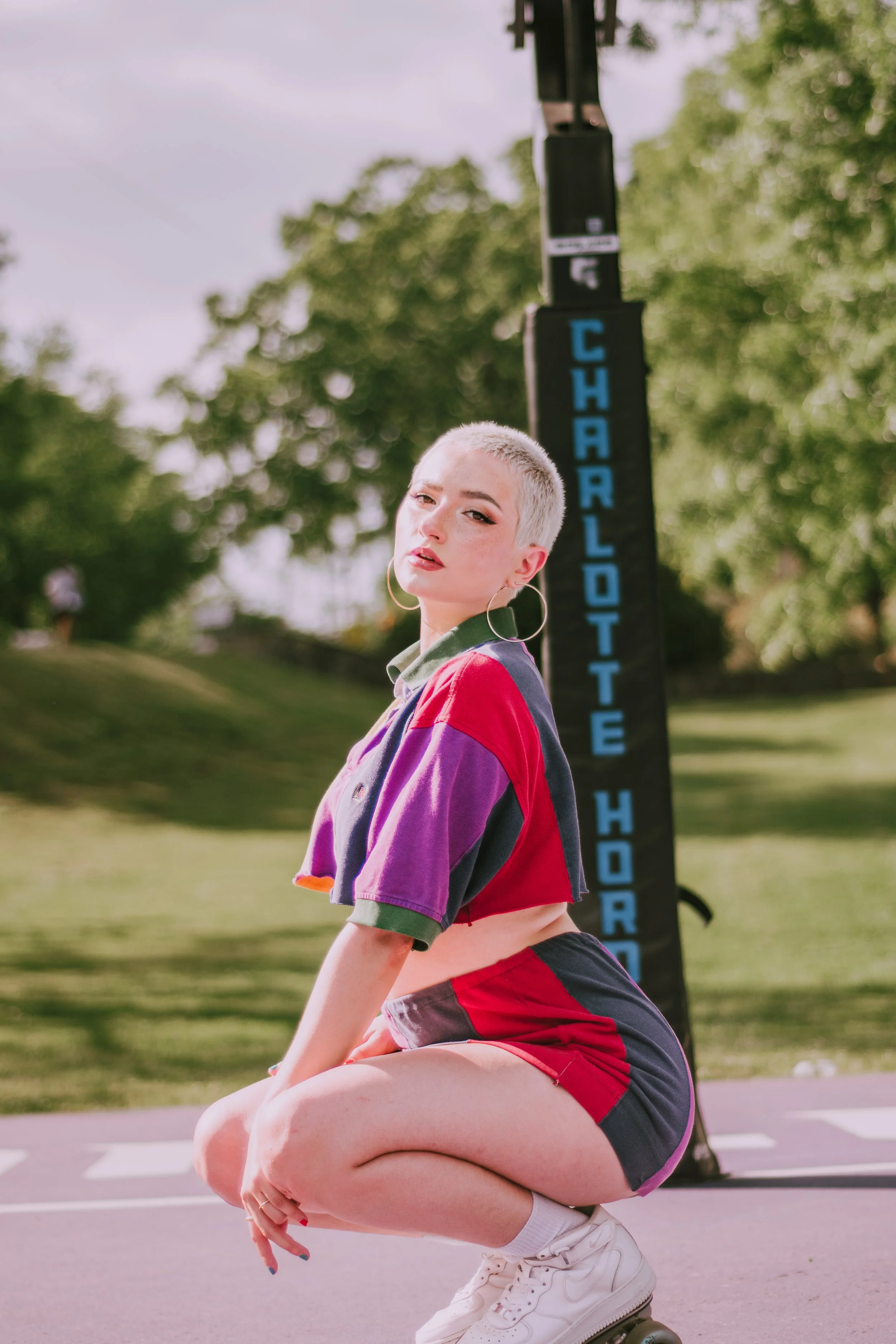 A young woman with short platinum blonde hair and hoop earrings is squatting on roller skates on a sidewalk. She is wearing a colorful athletic jacket and red shorts. Behind her is a street sign that says 'CHARLOTTE H' and a tree with green leaves.