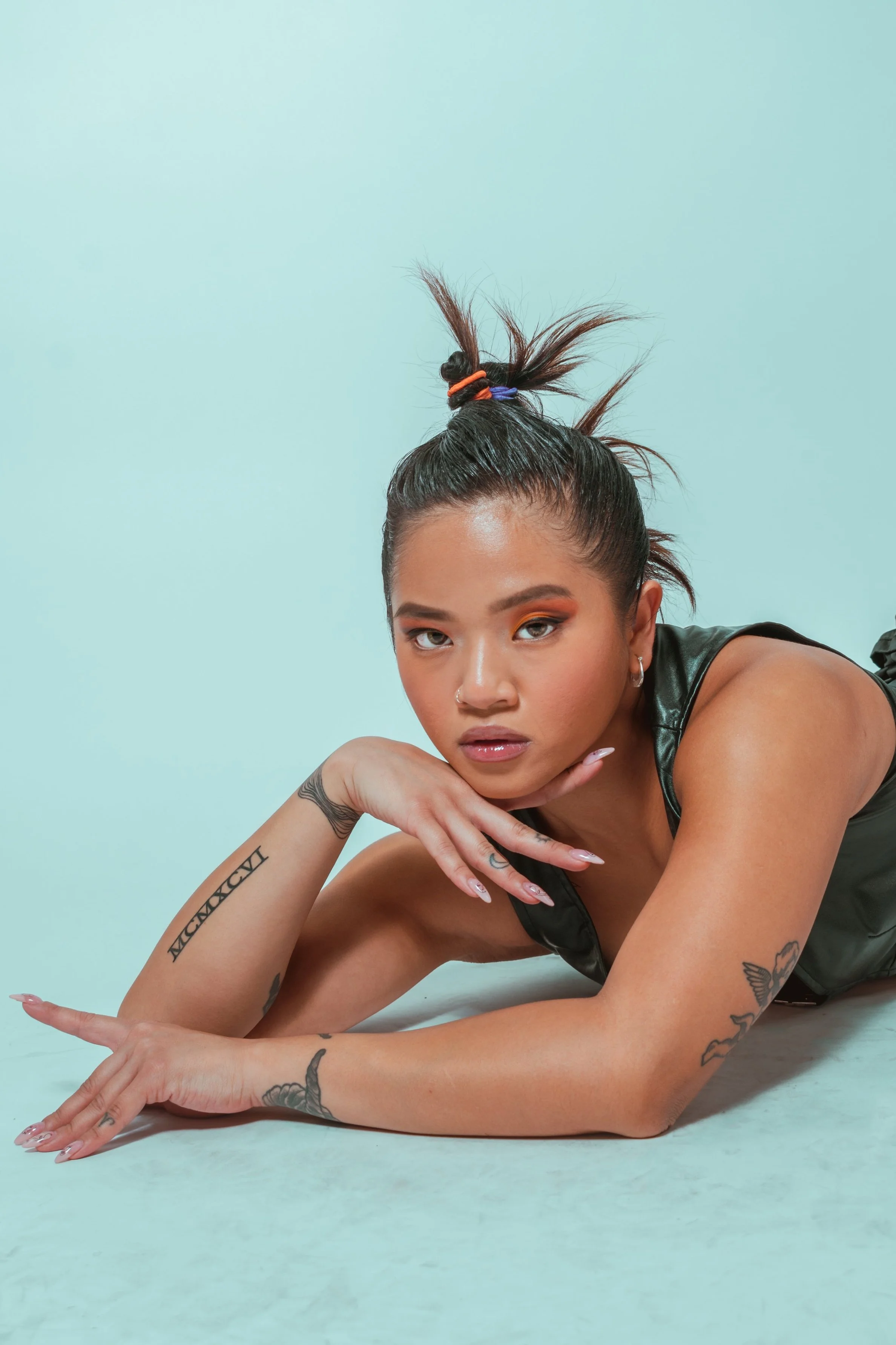 A young woman with tattoos and makeup resting her chin on her hand while lying on a light blue background.
