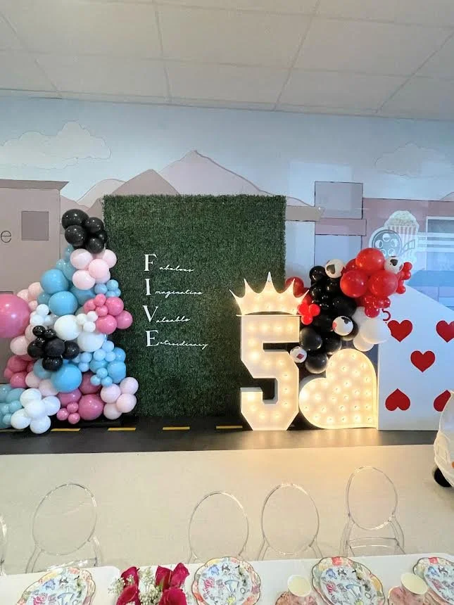 Birthday party decoration with a grass wall background, balloon arrangements, and a light-up number five with a crown. The word 'FIVE' and related words are displayed vertically. Plates and napkins are set on a table in the foreground.
