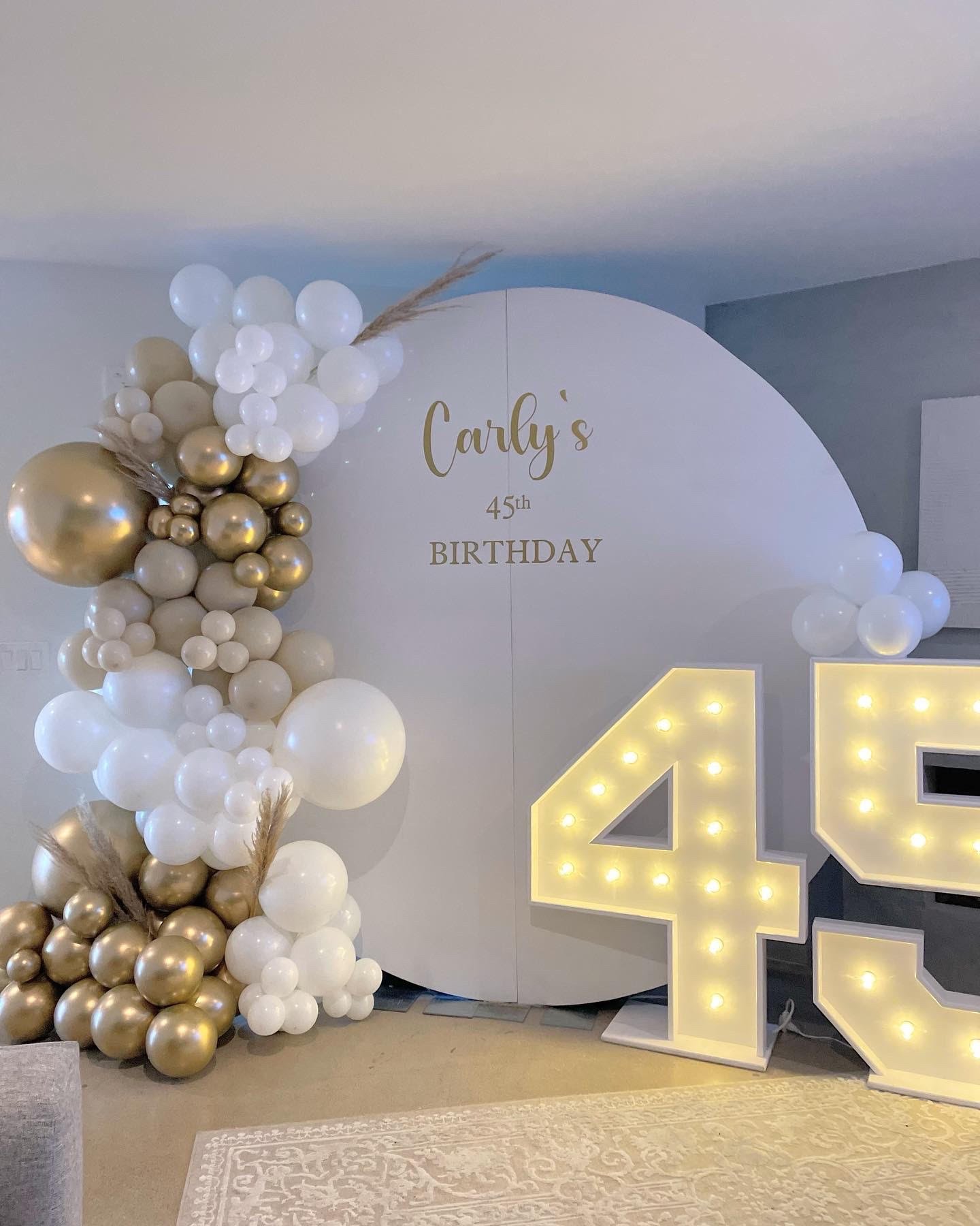 Birthday party decoration featuring a large white backdrop with "Carly's 45th Birthday" in gold lettering, surrounded by clusters of gold and white balloons. A large illuminated '45' is displayed next to the backdrop.