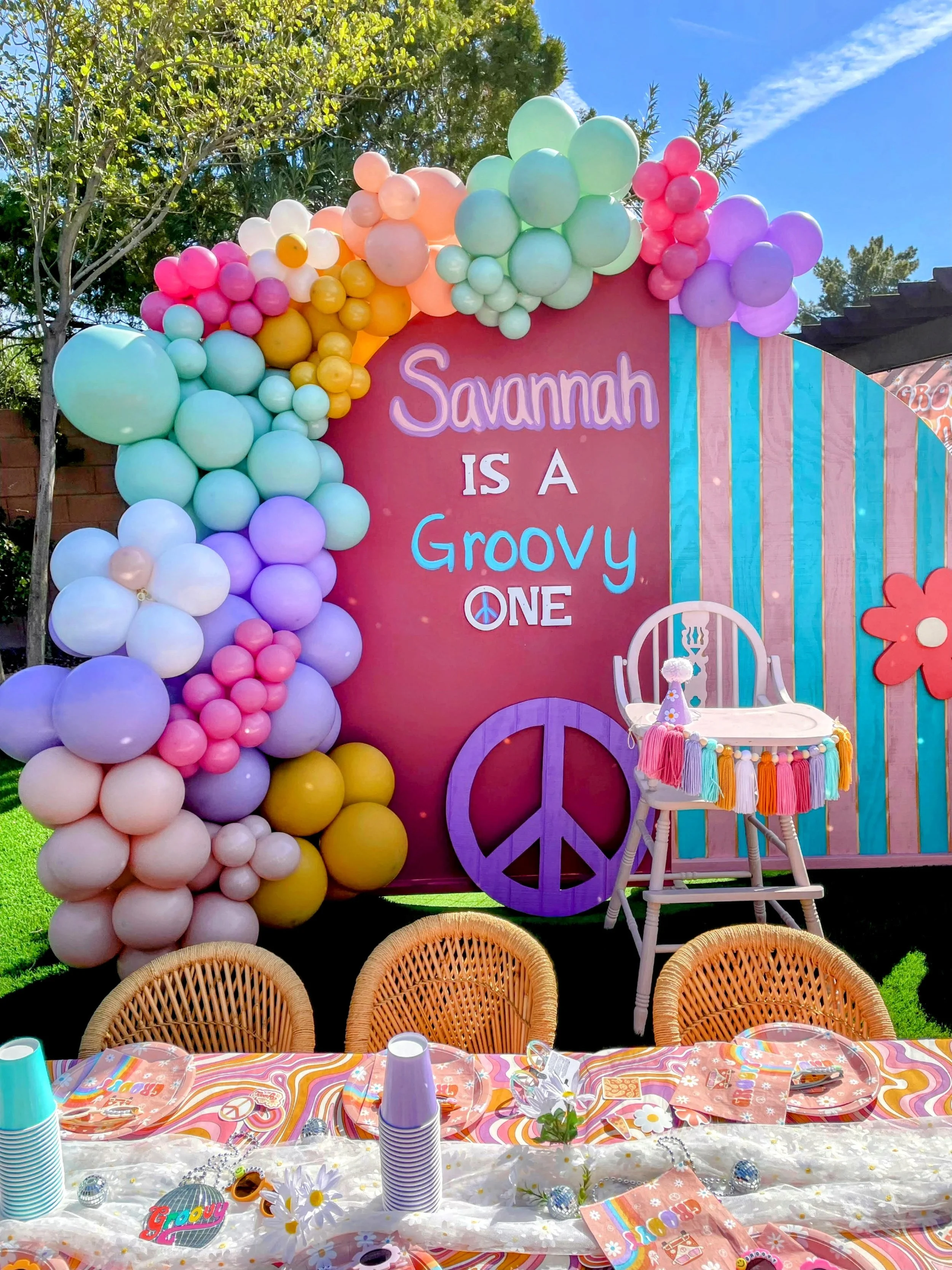 Birthday party setup with a colorful balloon arch and a sign reading "Savannah is a groovy one." A high chair is decorated with tassels, and a table is set with plates, cups, and a peace sign theme. The background includes trees and a blue sky.