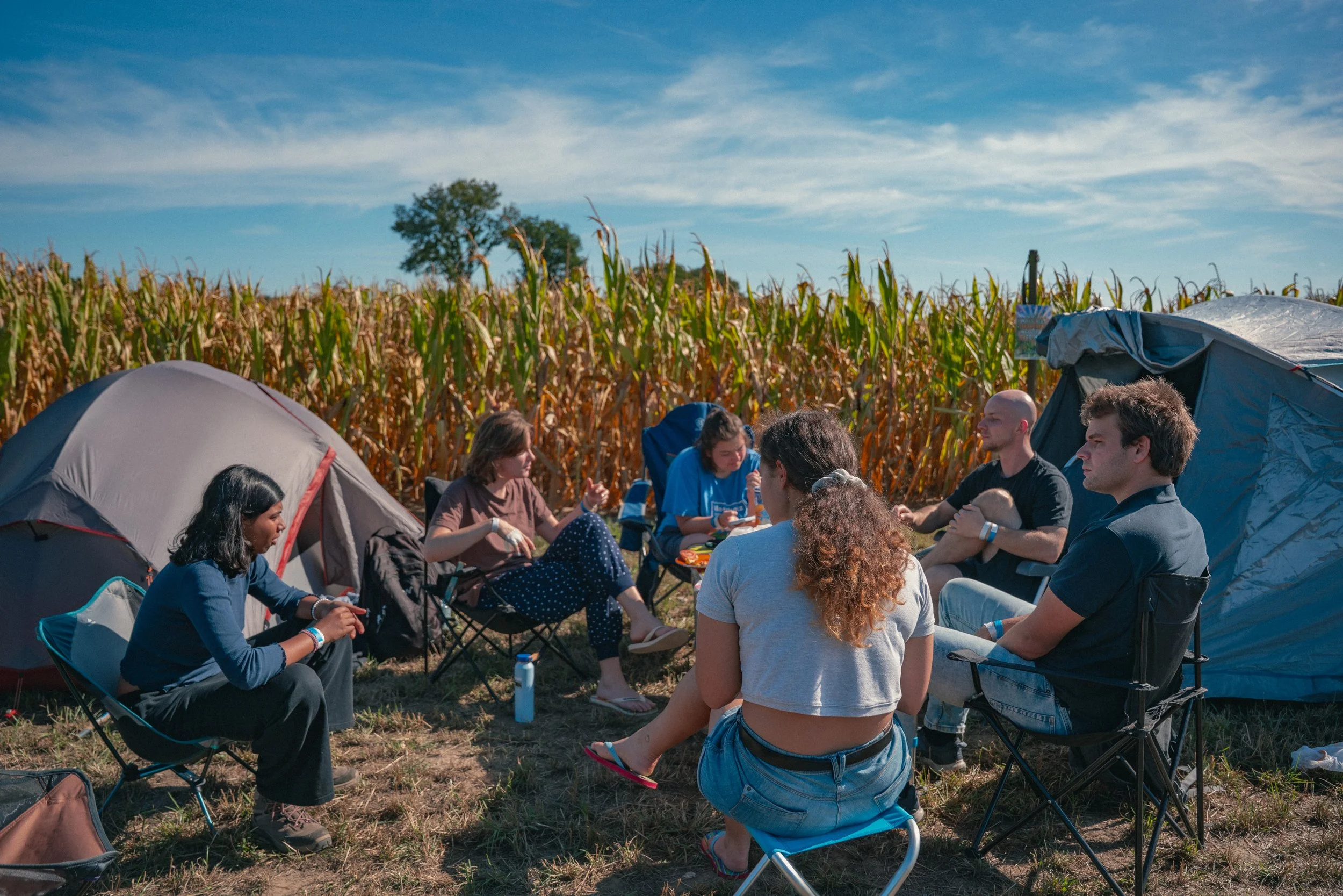 Mensen zitten in campingstoelen en tenten op een veld, met maïsvelden en een blauwe lucht op de achtergrond.