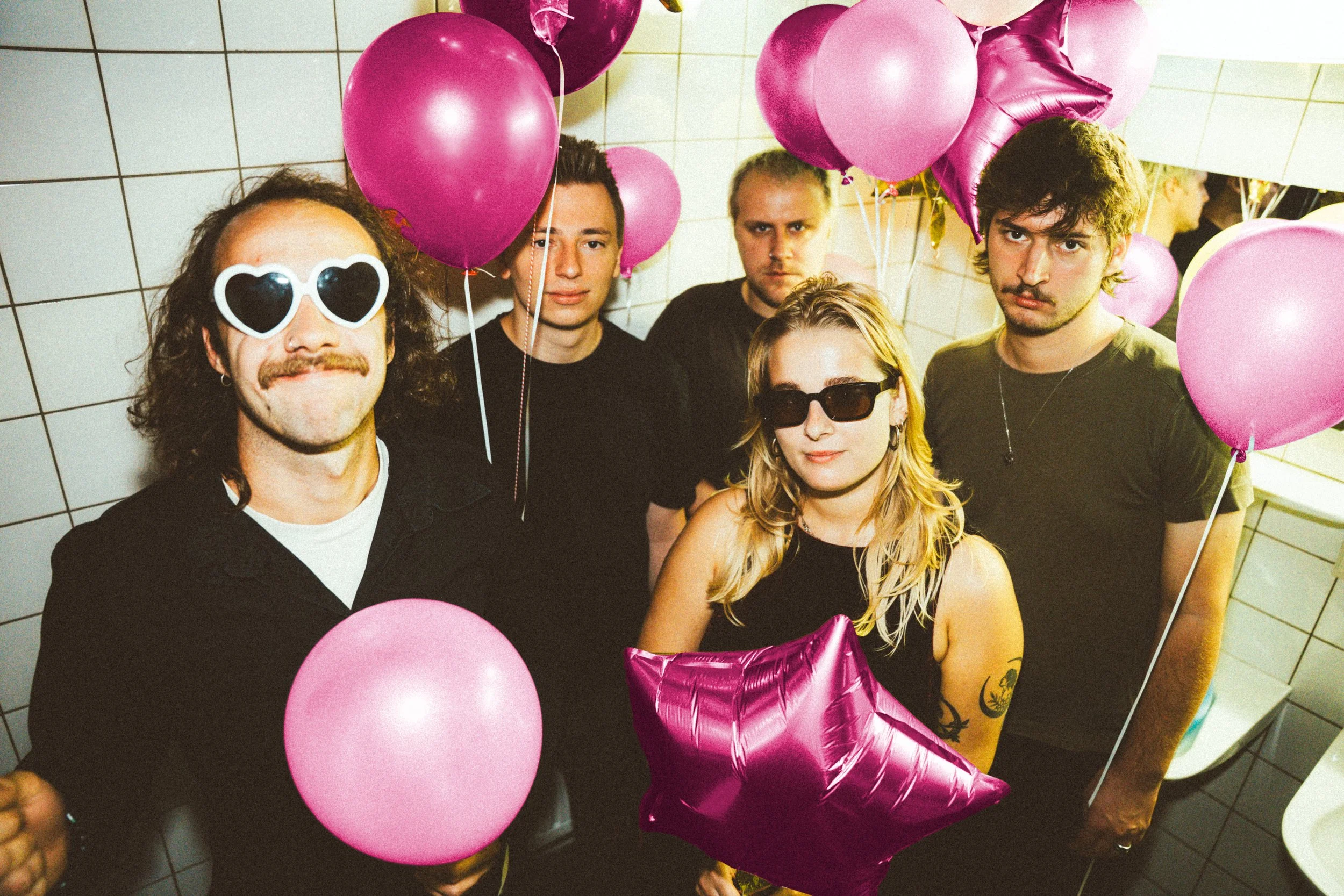 Vijf jongeren staan samen in een ruimte met witte tegels aan de muur, omringd door roze en paarse ballonnen, sommigen dragen zonnebrillen en feestkleding, en ze poseren voor een feest of feestje.