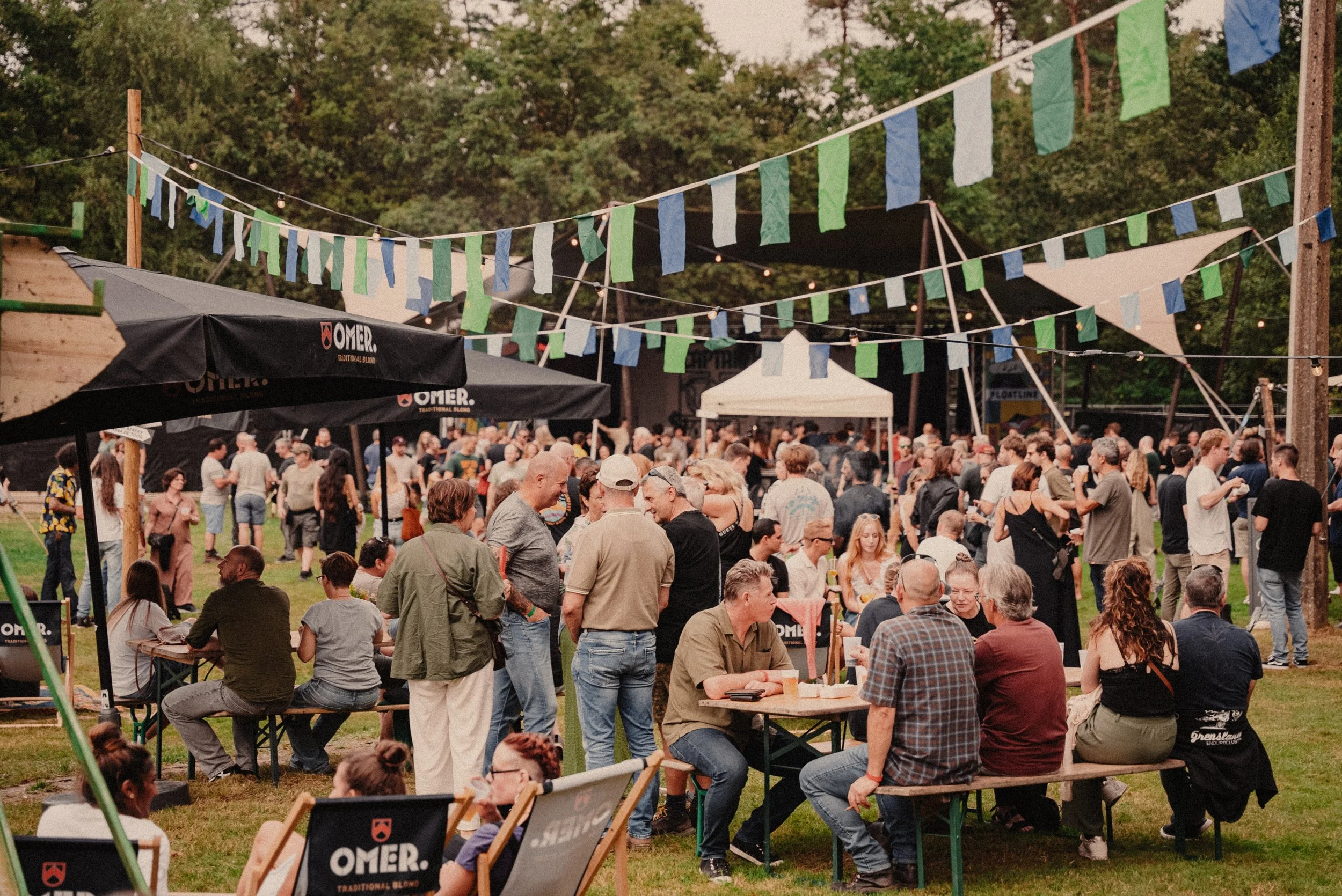 Een drukke enthousiaste menigte bij een outdoor festival met kleurrijke vlaggen en parasols, groene bomen op de achtergrond.