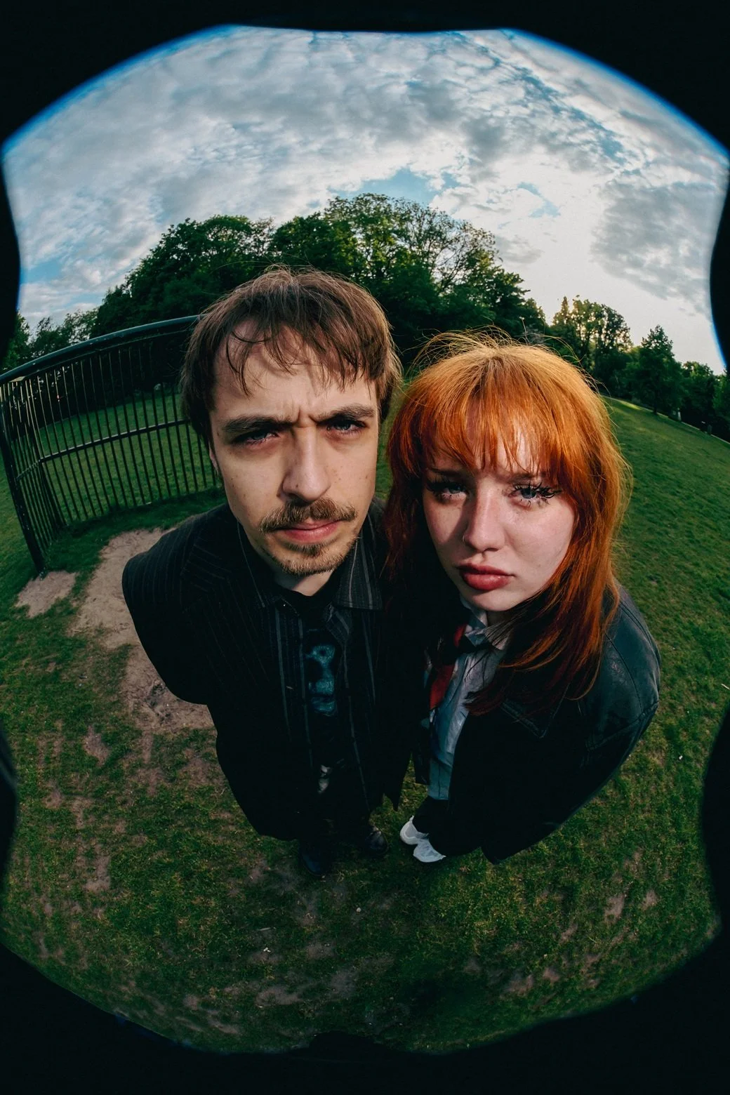 Tweedelige foto van een man en een vrouw die naar de camera kijken, genomen met een vislens. Beiden staan op een grasveld met bomen op de achtergrond, onder een bewolkte hemel.